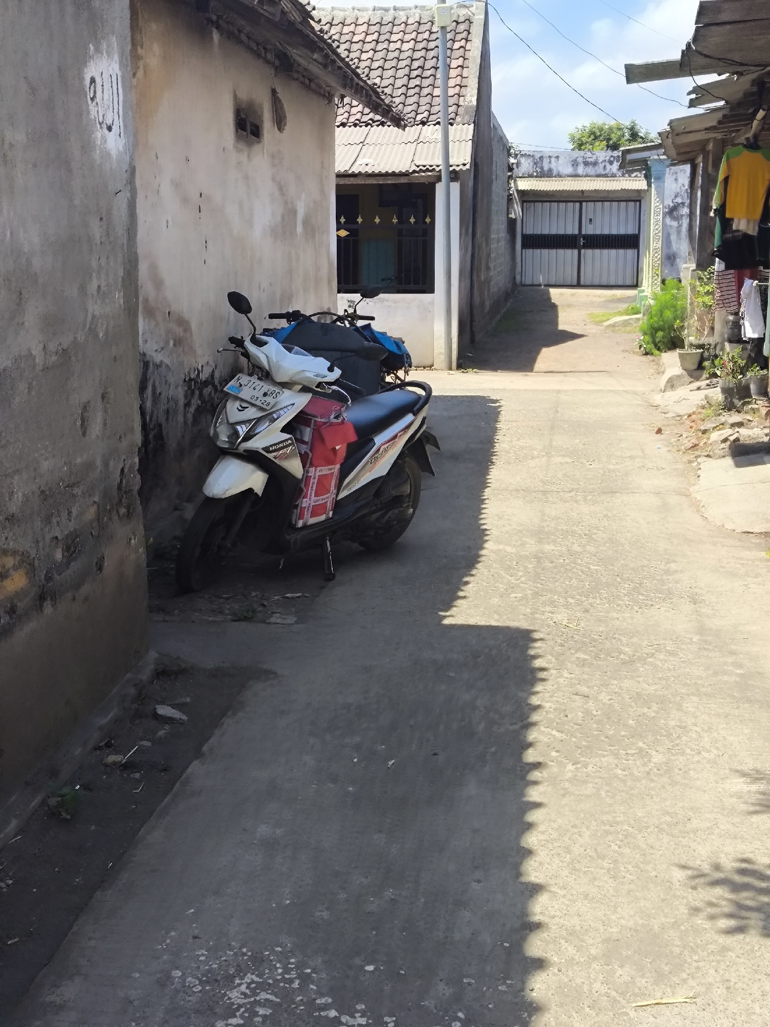 The image depicts a narrow, paved alleyway in Grati, Indonesia. On the left, a weathered, light-colored wall has Arabic script painted in black. Adjacent to this wall, a white and blue Honda scooter is parked. The scooter is equipped with a red carrier bag on its back. The alleyway continues into the background, leading towards a closed metal double gate. On the right side of the alley, a building has clothes hanging outside, including a yellow and green shirt and several other items. Potted plants are also visible along the right side. The sky above is blue with scattered white clouds. The scene is illuminated by sunlight, casting shadows on the ground.