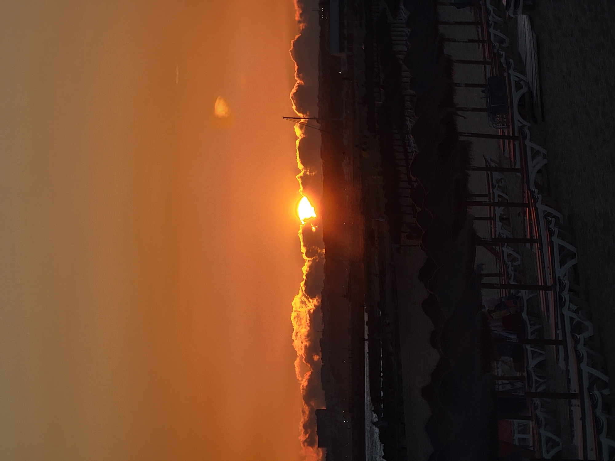 An outdoor shot captures a sunset over a beach in Quarteira, Portugal. The sky is a vibrant orange, with the sun partially obscured by a line of clouds that glow with the same warm hue. Below the sky, a silhouetted cityscape is visible, with buildings and a few lights. A body of water, possibly the ocean, is in the mid-ground, with the edge of a rocky shore in the foreground. The beach itself is lined with rows of empty beach umbrellas and lounge chairs, all in silhouette, suggesting the day is ending. The scene is tranquil, with no people or actions clearly depicted.
