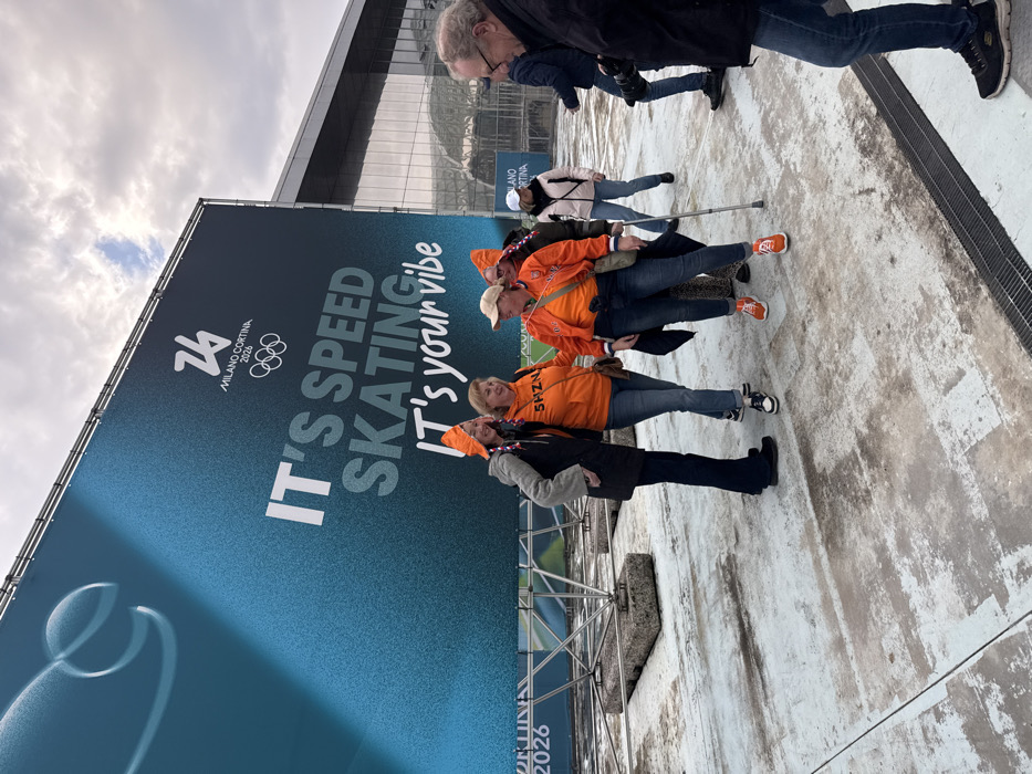 The image displays an outdoor scene in Rho, Italy, featuring a large promotional banner and several individuals. The dominant feature is a tall, rectangular banner in the left and center background, colored in a teal to dark blue gradient. It prominently displays "MILANO CORTINA 2026" with the Olympic rings logo, and the text "IT'S SPEED SKATING IT's your vibe." The banner is supported by a metal truss system with concrete blocks at its base.
In the mid-ground, a group of four women is posing for a photograph. Three of these women are distinctly dressed in orange shirts or jackets and orange cone-shaped hats. The leftmost woman wears a dark coat over a grey top and an orange hat. Next to her, a woman in an orange shirt with "SHZN" visible, blue jeans, and sneakers smiles at the camera. To her right, another woman in an orange jacket, orange top, blue jeans, orange sneakers, and an orange hat holds a grey crutch in her left hand while smiling at the camera. The fourth woman in this group is partially obscured.
To the right, a man wearing glasses, a dark jacket, and jeans holds a camera with a substantial lens, pointing it towards the posing group. Another man with a camera is visible in the mid-background. A woman in a light-colored jacket and dark pants, also using a crutch, is seen further back to the right of the main group.
The ground is a light-colored, possibly concrete or paved surface, exhibiting wear and darker stains. A metal grate is present in the immediate foreground. Beyond the banner, a modern building with a glass facade and a metallic roof structure is visible under a cloudy, overcast sky.