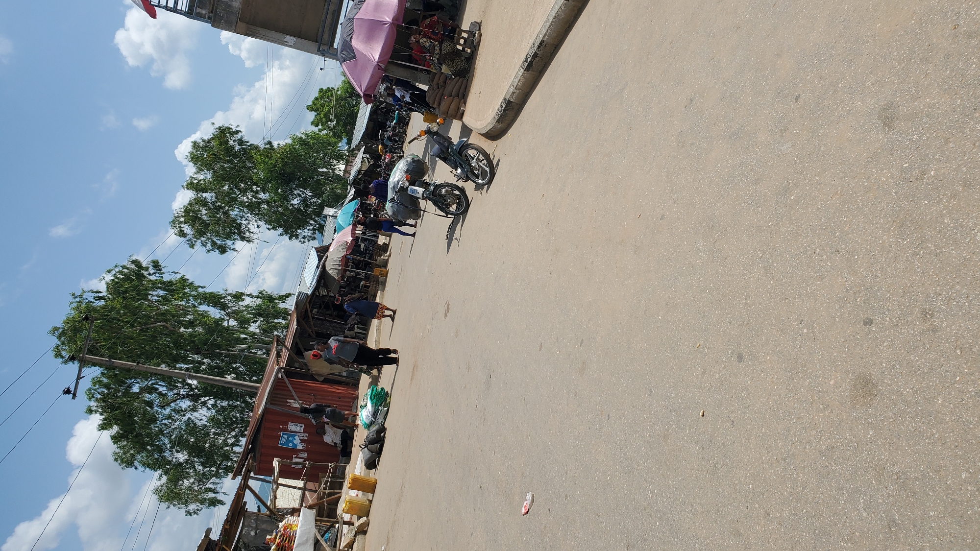 The image depicts a street scene in Jalingo, Nigeria, likely a marketplace, under a bright, partly cloudy sky. In the foreground, the asphalt road is visible, with a few scattered debris items. To the left, a section of market stalls with corrugated metal roofs and red shipping containers is set up. Several yellow plastic containers and dark bags are piled on the ground near the stalls. People are seen walking and standing in and around the market area.
Further into the scene, more market stalls are present, many covered by colorful umbrellas, indicating outdoor selling. A few motorcycles are parked on the side of the road, one of which is loaded with large bags. People are interacting within the market, with some standing, others sitting, and some walking between the stalls. In the background, tall trees with lush green foliage are prominent, and a utility pole with wires extends upwards. On the right side of the image, a concrete structure, possibly a bridge or overpass, is partially visible. The overall impression is of a busy, everyday commercial environment in Jalingo.