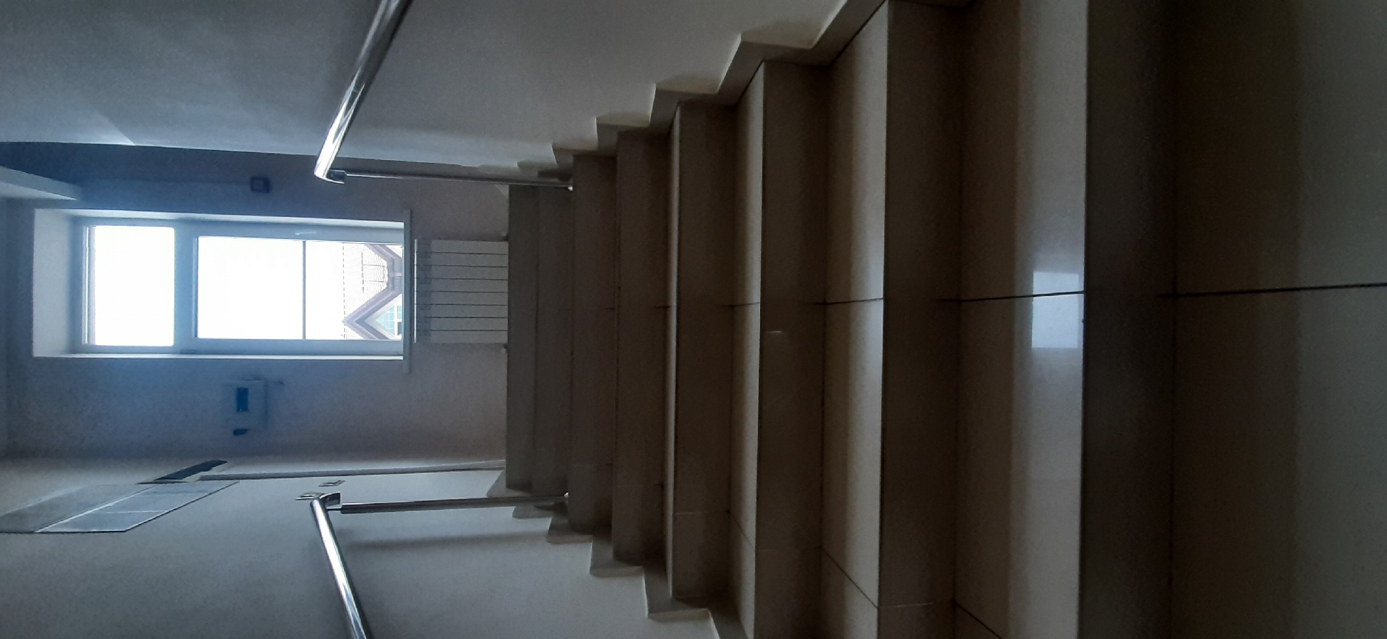 This is an indoor, eye-level shot of a modern staircase in Yessentuki, Russia. The scene is illuminated by natural light streaming through a large window on the left side of the frame. The window has two panes and looks out onto a building with a distinctive geometric roofline, suggesting an urban or suburban setting. Below the window, a white radiator is mounted on the wall.
The staircase itself is made of light-colored, smooth material, possibly stone or tile, with a polished finish. The steps have a tiered design, creating a sense of depth and perspective as they ascend. A metallic handrail runs along the side of the stairs, providing a sleek contrast to the neutral tones of the steps and walls. The lighting is somewhat dim, casting shadows that accentuate the architectural features of the staircase.
To the left of the stairs, a light gray wall is visible, with a small, blue rectangular object mounted on it, possibly a utility box or intercom. The floor in front of the stairs appears to be tiled or covered with a similar smooth material, with some rectangular metallic plates set into it. The overall impression is one of clean lines and functional design, typical of contemporary architecture. There is no visible text in the image. The time of day is suggested to be daytime due to the natural light.