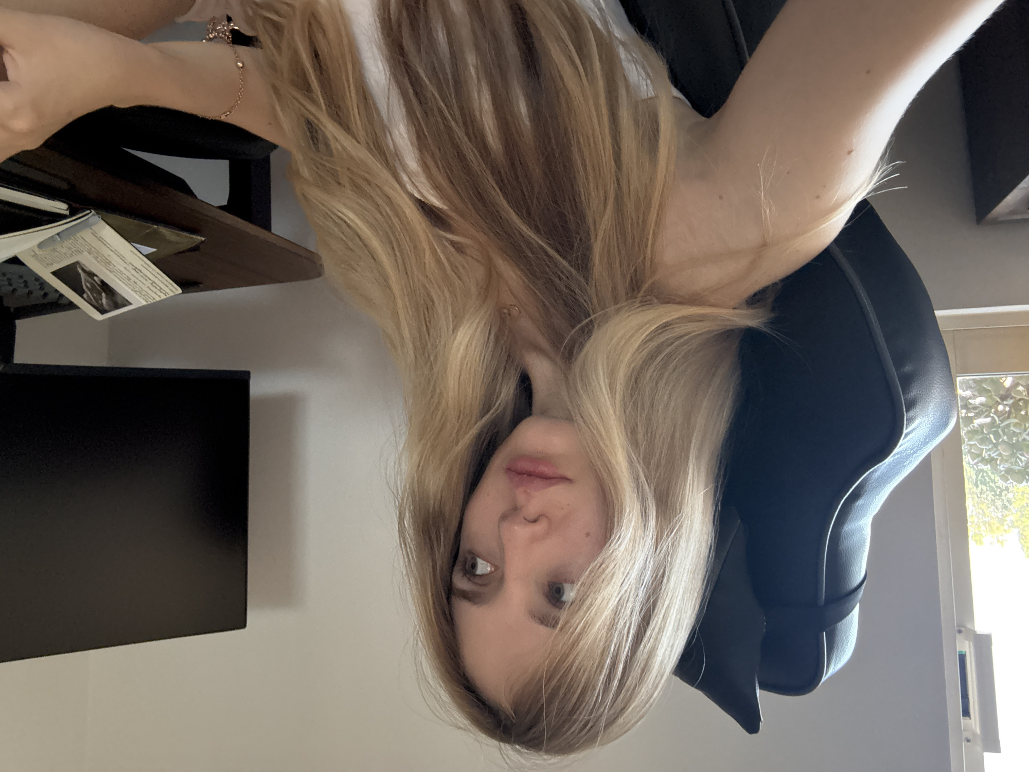 The image displays a young woman indoors, in what appears to be a home office or living space in Mexico City, Mexico. She is captured from an unusual, inverted angle, with her long, wavy blonde hair flowing downwards towards the top of the frame. Her light-colored eyes gaze directly at the camera with a neutral, slightly contemplative expression. She has light skin and is wearing a light-colored top, possibly white.
She is seated in a black, high-backed office or gaming chair, parts of which are visible supporting her. On her left wrist, a delicate, gold-toned bracelet with a small charm is noticeable. Entangled in her hair near her neck, a small, circular, gold-colored pendant or earring is visible.
To her left, a dark wooden desk or shelf holds several papers. One document clearly features a small, framed image of a person (possibly a drawing or sketch) accompanied by small, illegible text columns. Below the desk, a dark, rectangular object, likely a computer monitor or television screen, is partially visible. To her right, a window allows bright natural light to stream in, revealing lush green foliage outside, indicating it is daytime. The room features light-colored walls. The image captures a casual, self-portrait-like moment.