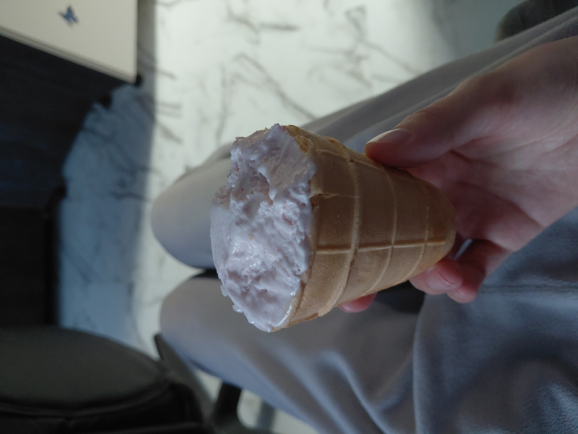 The image shows a close-up perspective of a person's hand holding a partially eaten ice cream cone indoors. The ice cream is a light pink color, likely strawberry or raspberry flavored, and appears slightly melted, dripping a little at the bottom. It is held in a classic waffle cone with a visible grid pattern. The person's hand is fair-skinned, with visible fingers and a thumb supporting the cone.
In the background, there is a light-colored surface with faint, wavy patterns, resembling marble or a textured wall. To the upper left, a blurry dark blue shape is visible, and on a white surface, there's a small, distinct blue butterfly shape. The person appears to be wearing soft, light gray clothing, possibly a sweatshirt or loungewear, which is visible around the hand and in the foreground. Another dark, indistinct object, possibly part of a chair or furniture, is visible in the lower-left corner. The overall lighting suggests an indoor setting. No text is visible in the image. The location context provided is Nadym, Russia.