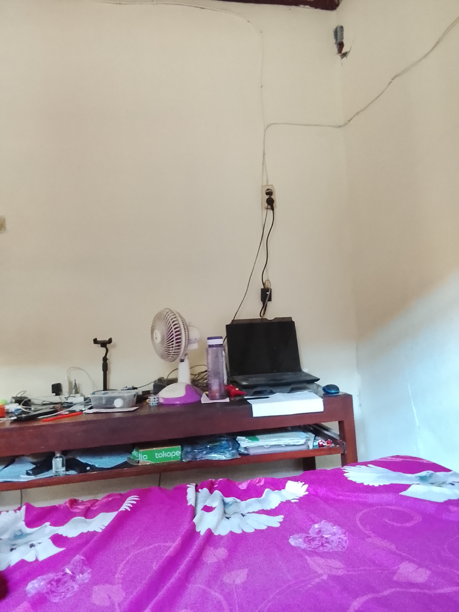 The image depicts a cluttered wooden desk against a light-colored wall in what appears to be a bedroom or study. A laptop is open on the right side of the desk, plugged into an electrical outlet on the wall above. To the left of the laptop, there are various items including a water bottle, a desk fan, a phone stand, and a container of items. Wires and cables are visible on the desk and running along the wall. Below the desk, there is a shelf with more items, including a green box with text that appears to be "jia tokopē". The foreground of the image is dominated by a bright pink fabric with a white bird pattern, suggesting it is a bedspread or blanket. The overall impression is of a lived-in, somewhat disorganized personal space. The location is identified as Karangklesem, Indonesia, based on the provided context.