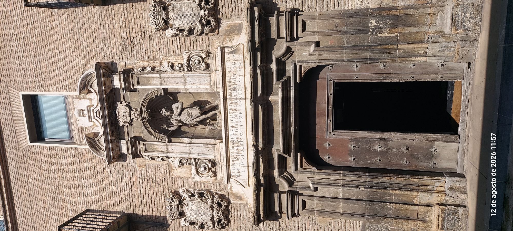 The image shows the ornate stone facade of a building with a large, weathered wooden double door opening into darkness. Above the doorway, a sculpted niche contains a statue of a bearded male figure holding a staff. To the left and right of the niche are carved coats of arms. A stone inscription is visible below the niche, reading "ESTE COLEGIO DE S.THOME BAP.STA FUNDARON LOS SEÑORES DVMANT Y BAPTISTA DE DVMANT Y ALDEXO ANNO DOMINI MDCCXLVIII ET SEÑORES DON M. RRIZ SVMVCER ANNO DE 1734". The building is constructed of brick with stone architectural details. The timestamp "12 de agosto de 2026 11:57" is visible in the bottom left corner. This facade is located on the Collegio de S. Tomás de Pamplona, Spain.