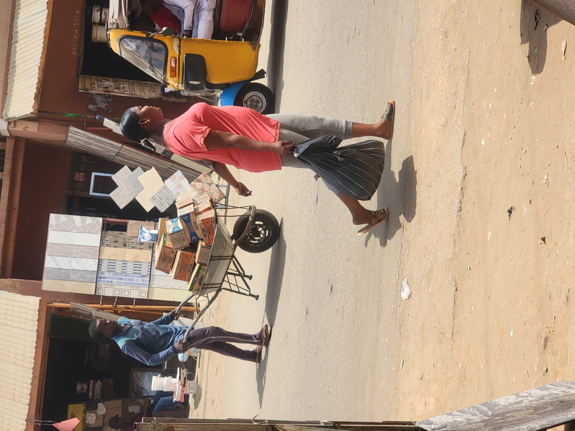 A daytime street scene in Maiburiji, Nigeria shows a woman in a pink top and grey leggings walking on a light tan paved road. She is carrying a striped plastic bag and wearing sandals. A man in a blue shirt and dark pants is pulling a metal cart loaded with boxes and tiles. A yellow three-wheeled vehicle is parked nearby. Buildings with various materials and awnings line the street.