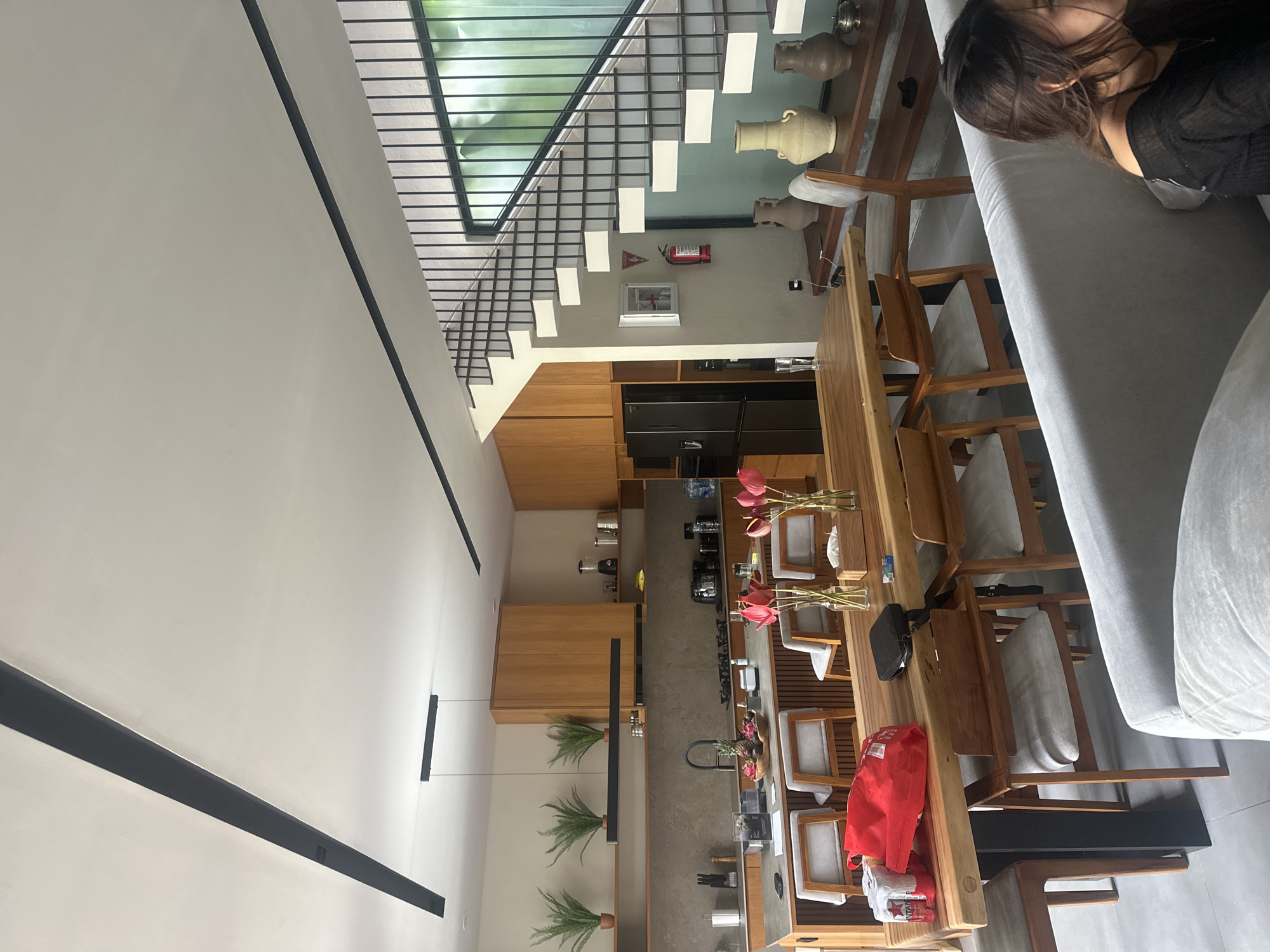 The image shows an interior view of a space that appears to be a modern house or apartment in North Kuta, Indonesia. A woman is seen lying on a light grey sofa. The living area extends into a dining area with a long wooden table and chairs. The kitchen area is beyond, featuring wooden cabinets and a dark refrigerator. The decor includes plants and what appears to be a fire extinguisher near a staircase that is partially visible. The overall design is minimalist and modern.
