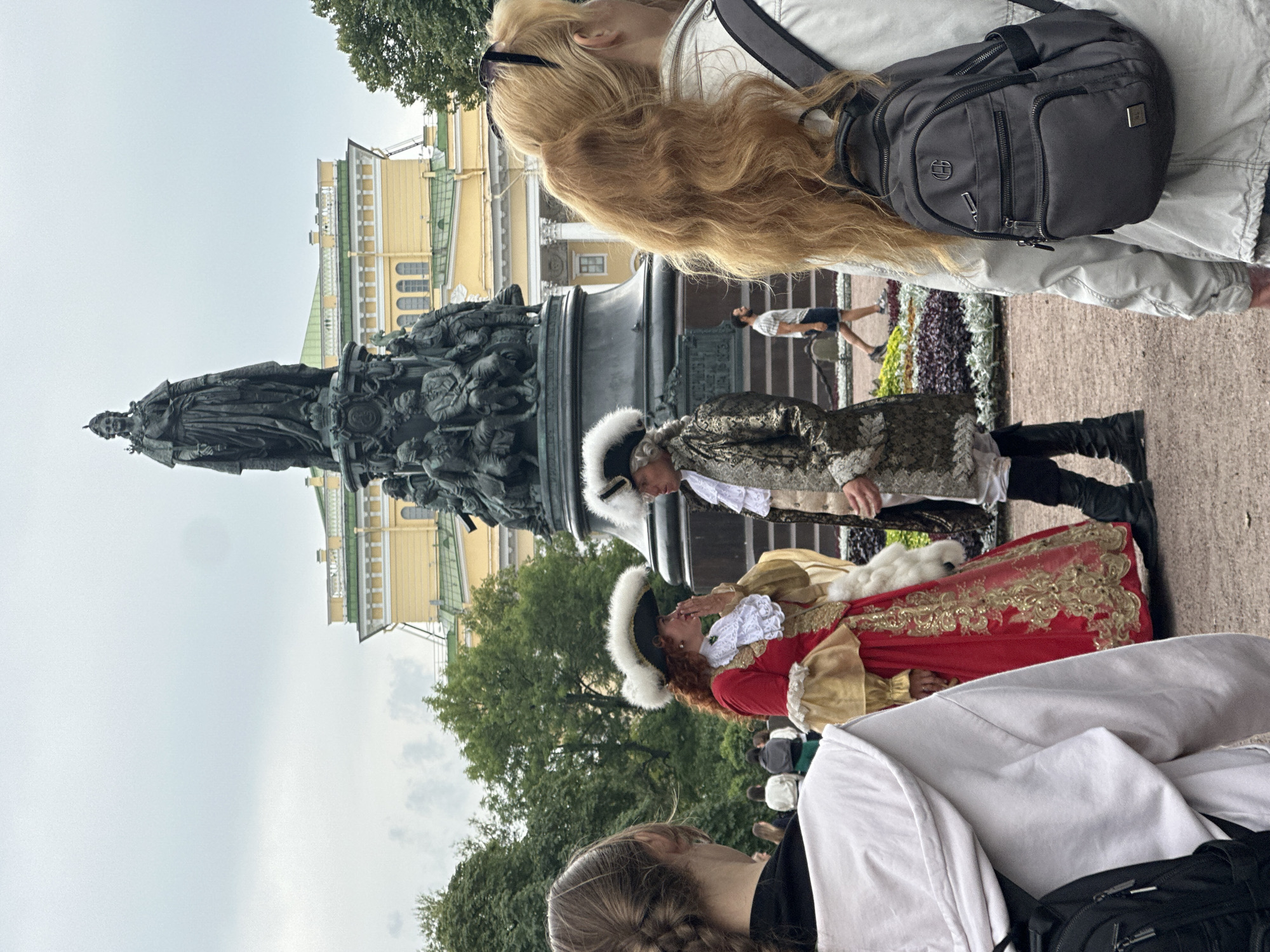 In Saint Petersburg, Russia, on a cloudy day, a monument to Catherine the Great stands prominently. The monument features a large statue of the empress atop an ornate pedestal adorned with smaller figures. In front of the monument, two individuals dressed in historical costumes, a woman in a red and gold gown with a large feathered hat and a man in a patterned coat and feathered hat, appear to be interacting. They are surrounded by lush greenery and flowerbeds. Several other individuals, dressed in modern clothing, are also present in the scene, some in the foreground and others in the background, including a person with red hair wearing a backpack. The background also shows a yellow building with green accents. The overall scene suggests a public space, possibly a park or a square, where historical reenactments or events might be taking place.