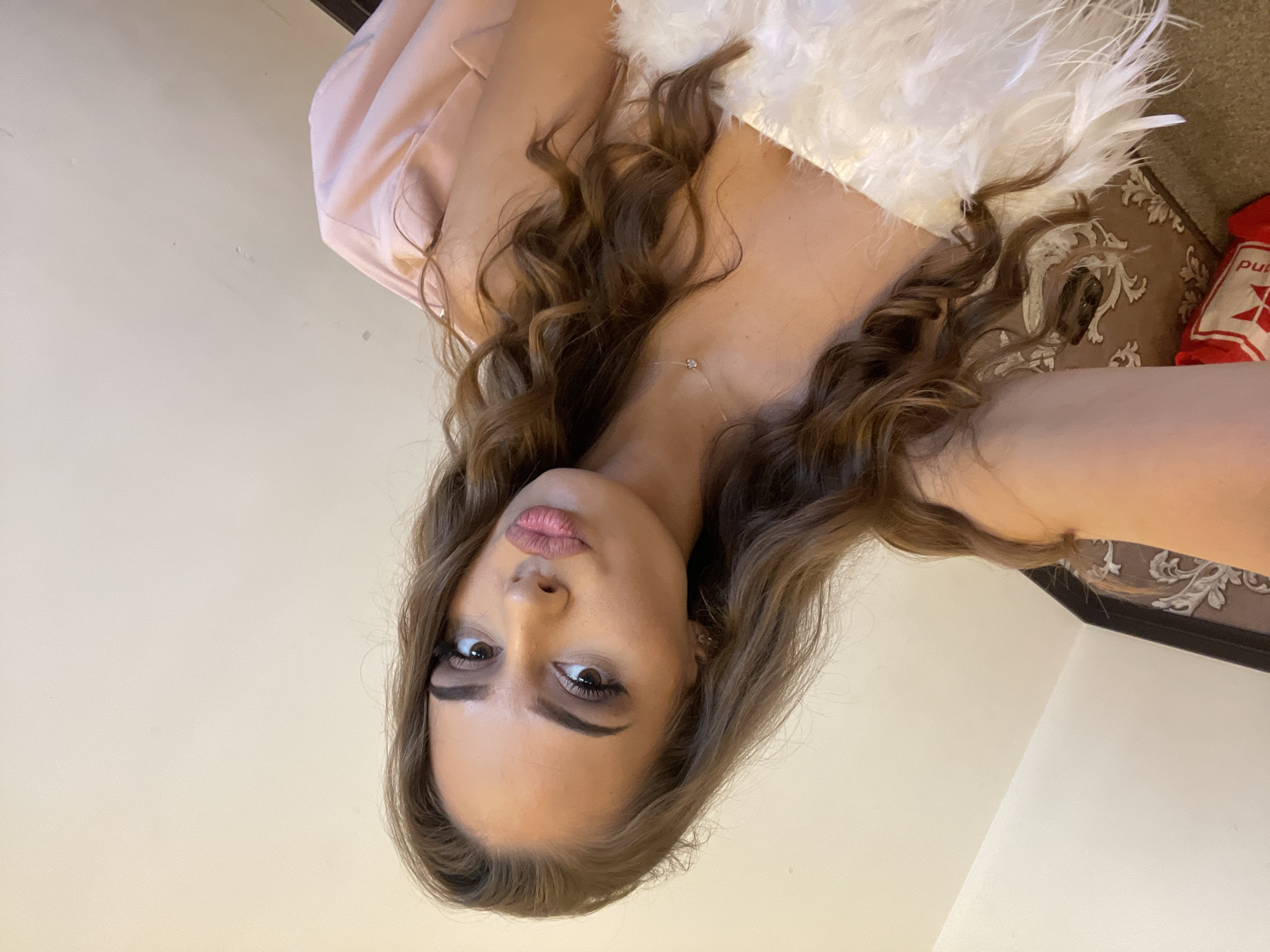 A woman with long, wavy brown hair is shown in a close-up, angled selfie. She has fair skin and is wearing makeup, including dark eyeliner and lipstick. She is wearing a light pink blazer and a white, feathered garment that obscures part of her upper body. A thin necklace is visible around her neck. The background includes a light-colored wall and a patterned brown cushion or surface near a red bag. The location is not discernible from the image.