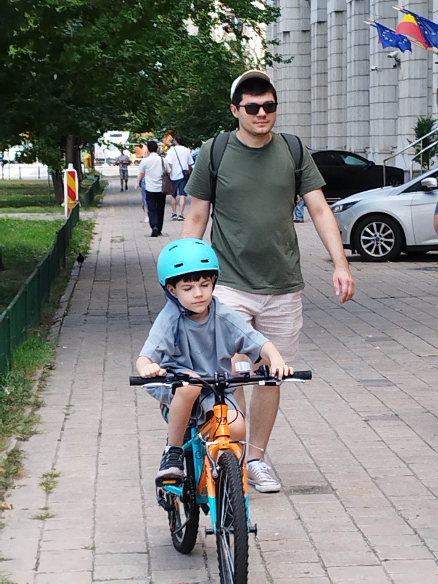In Bucharest, Romania, a young boy wearing a blue helmet rides a bicycle on a paved sidewalk. An adult male, wearing a green t-shirt, shorts, sunglasses, and a white cap, walks beside the boy. The background features trees, other pedestrians, parked cars, and a building with columns and flags.