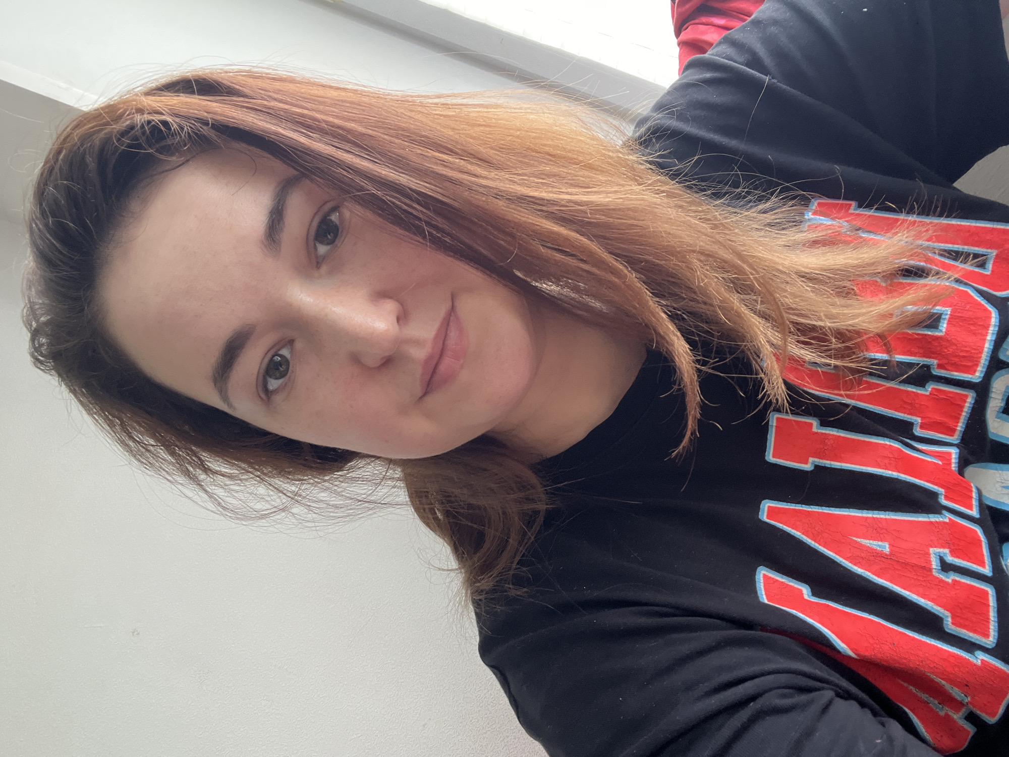 A woman with reddish-brown hair and freckles is looking at the camera. She is wearing a black t-shirt with red and blue lettering. The background is a plain white wall and a window with natural light coming through. There is no indication of the city of Chelyabinsk, Russia, or any other specific location.