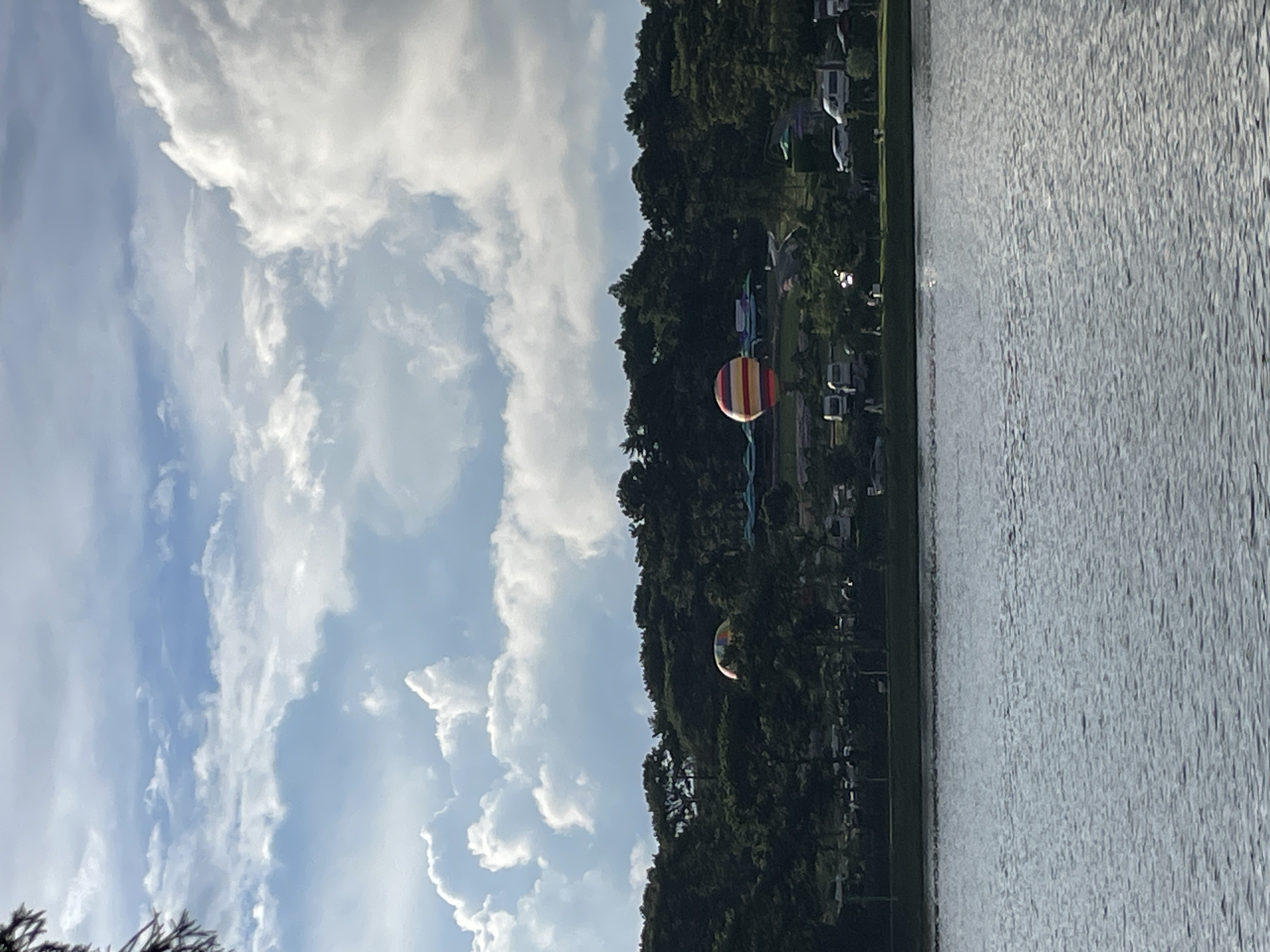 This image depicts a scenic view of a lake or body of water with a tree-covered hillside in the background. The sky is filled with scattered white clouds against a blue backdrop, suggesting a pleasant day. Two hot air balloons are visible. One, larger and closer, is painted with horizontal red and yellow stripes. The other, smaller and further away, appears to be a more muted color. The hillside is densely wooded, and several small structures, possibly houses or campsites, are nestled among the trees. The water's surface is textured with ripples, reflecting the light. The overall scene evokes a sense of tranquility and outdoor recreation. The presence of the hot air balloons suggests an event or festival might be taking place. The lighting indicates it is likely daytime, possibly late afternoon given the softness of the light.