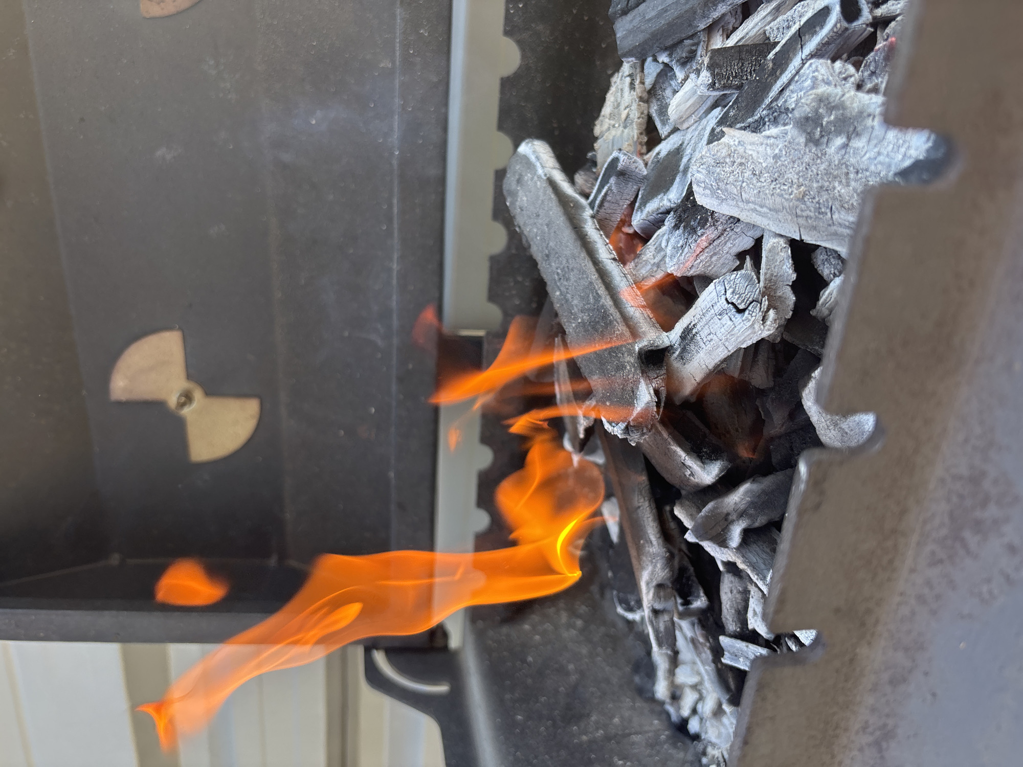The image depicts burning charcoal inside a grill. The fire is visible, with flames extending to the left side of the grill. Charcoal pieces, partially consumed, are inside the grill. A metal airflow control dial is visible on the left. The location and city of Chaikovsky, Russia, cannot be determined from the image.