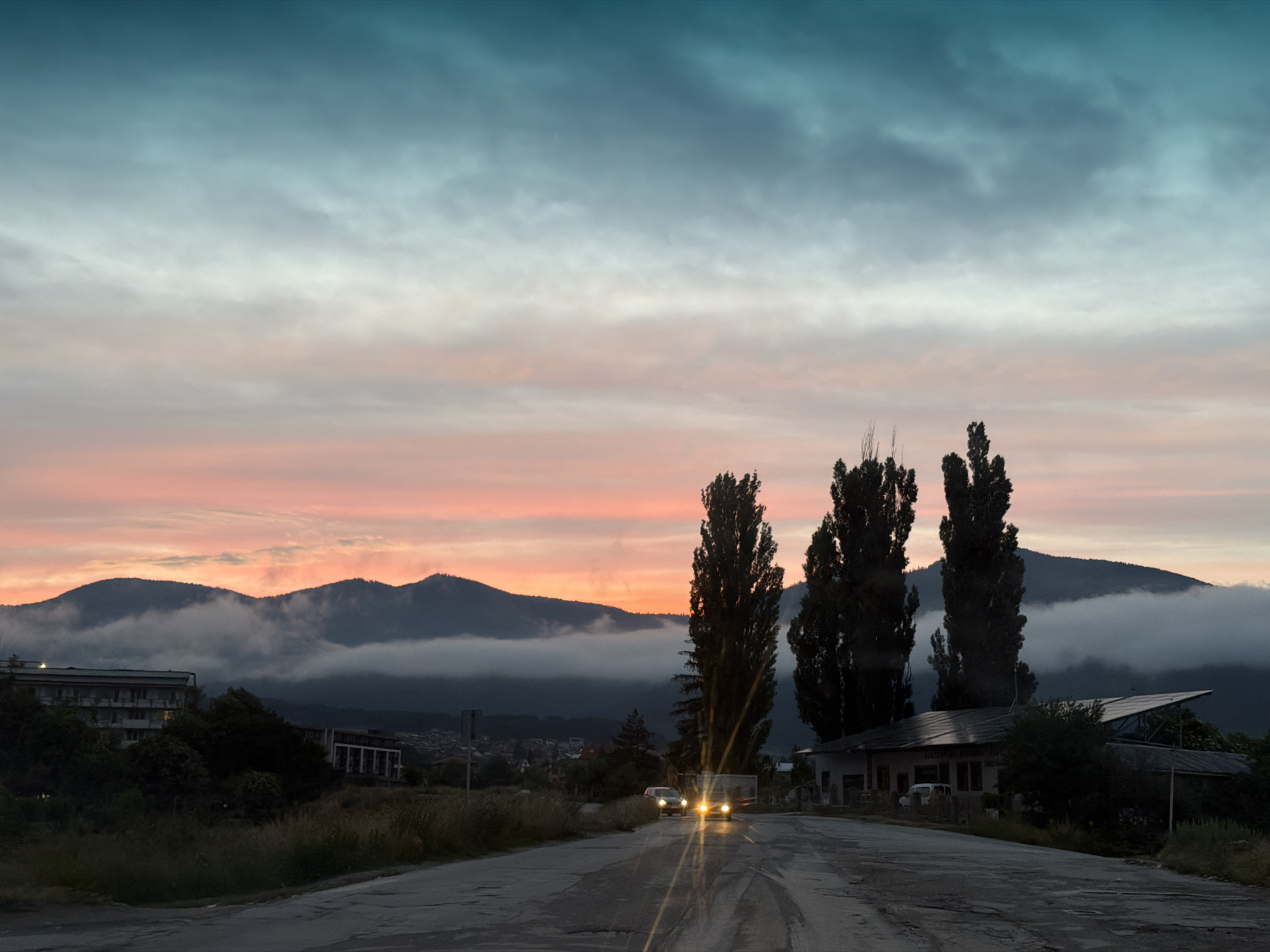 A long, winding, wet asphalt road leads into a landscape dominated by a dramatic sky at dusk, featuring hues of teal fading into soft pinks and oranges. Mountains shrouded in fog form the backdrop, their peaks breaking through the mist. On the left, a long, multi-story building with many windows sits amongst dark green trees. To the right of the road, a low-lying building with a slanted roof, possibly adorned with solar panels, is visible, surrounded by vegetation. Several tall, slender trees stand in the middle ground, their dark silhouettes contrasting with the lighter sky. Two cars with their headlights on are approaching on the wet road, their bright lights reflecting on the surface. The overall scene suggests a tranquil yet dynamic twilight setting in Velingrad, Bulgaria, with an emphasis on the interplay of natural light and atmospheric conditions.