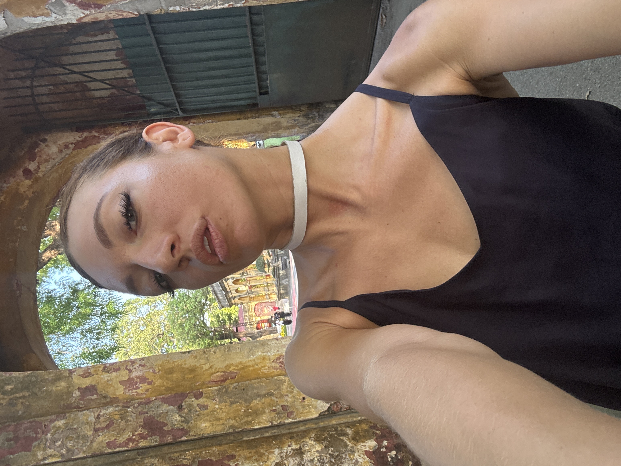 A selfie shows a woman with pale skin and brown hair, wearing a black top and white choker necklace, looking into the camera. She is positioned within an old, arched stone or concrete structure with peeling paint. Through the archway is a view of a building with intricate architectural details, trees, and other people, indicating an outdoor location. The location is in the city of Hà Nội, Vietnam.
