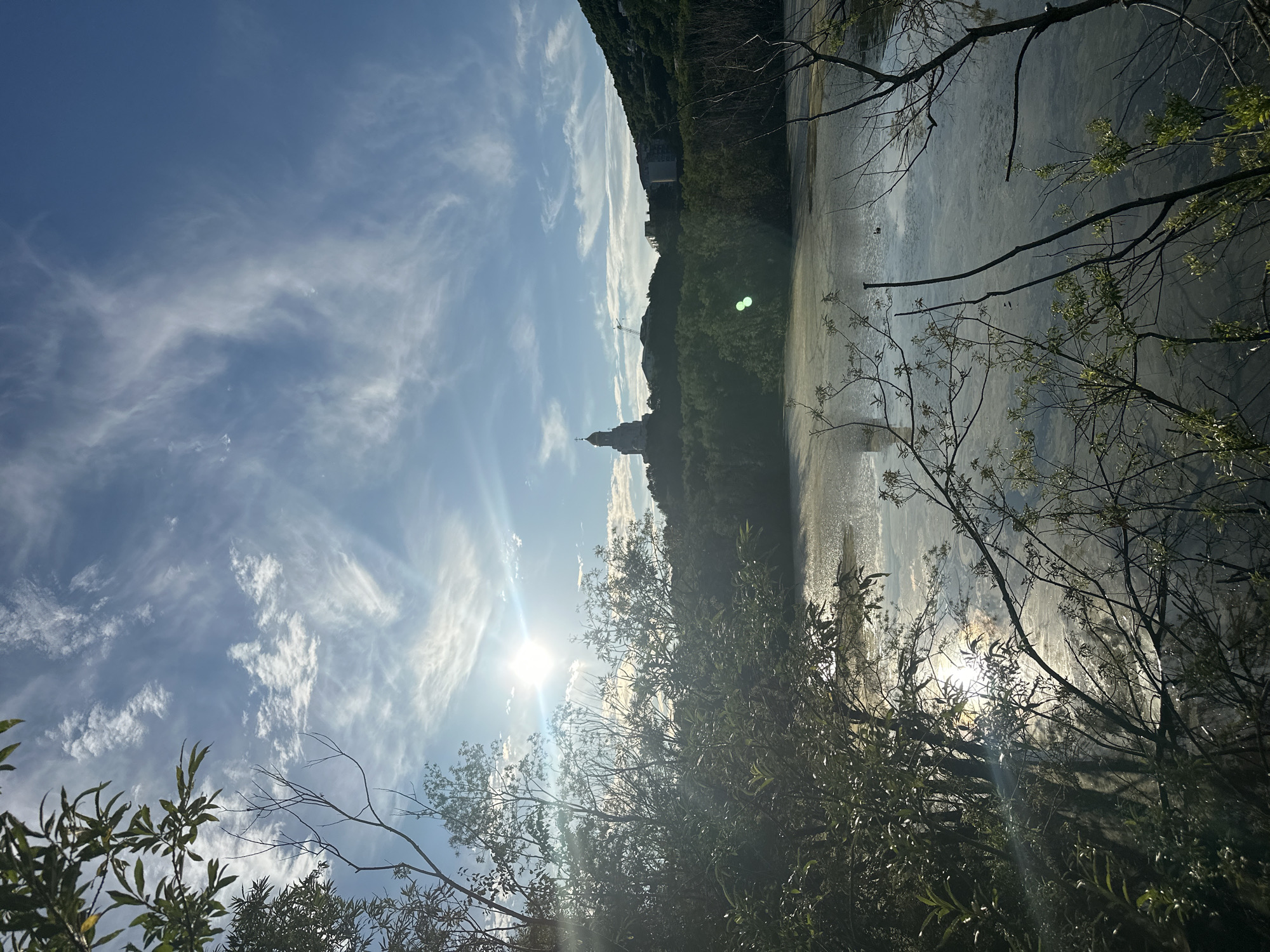 The image captures a serene landscape with a body of water in the foreground, bordered by lush greenery and silhouetted trees. The sun, positioned in the upper left quadrant, casts a bright glare, reflecting off the water's surface and illuminating wispy clouds in a clear blue sky. In the distance, a prominent building with a domed tower stands atop a hill, surrounded by other structures and dense vegetation. The overall scene evokes a peaceful, natural setting with architectural elements visible in the background. There are no people or animals clearly discernible in the image.