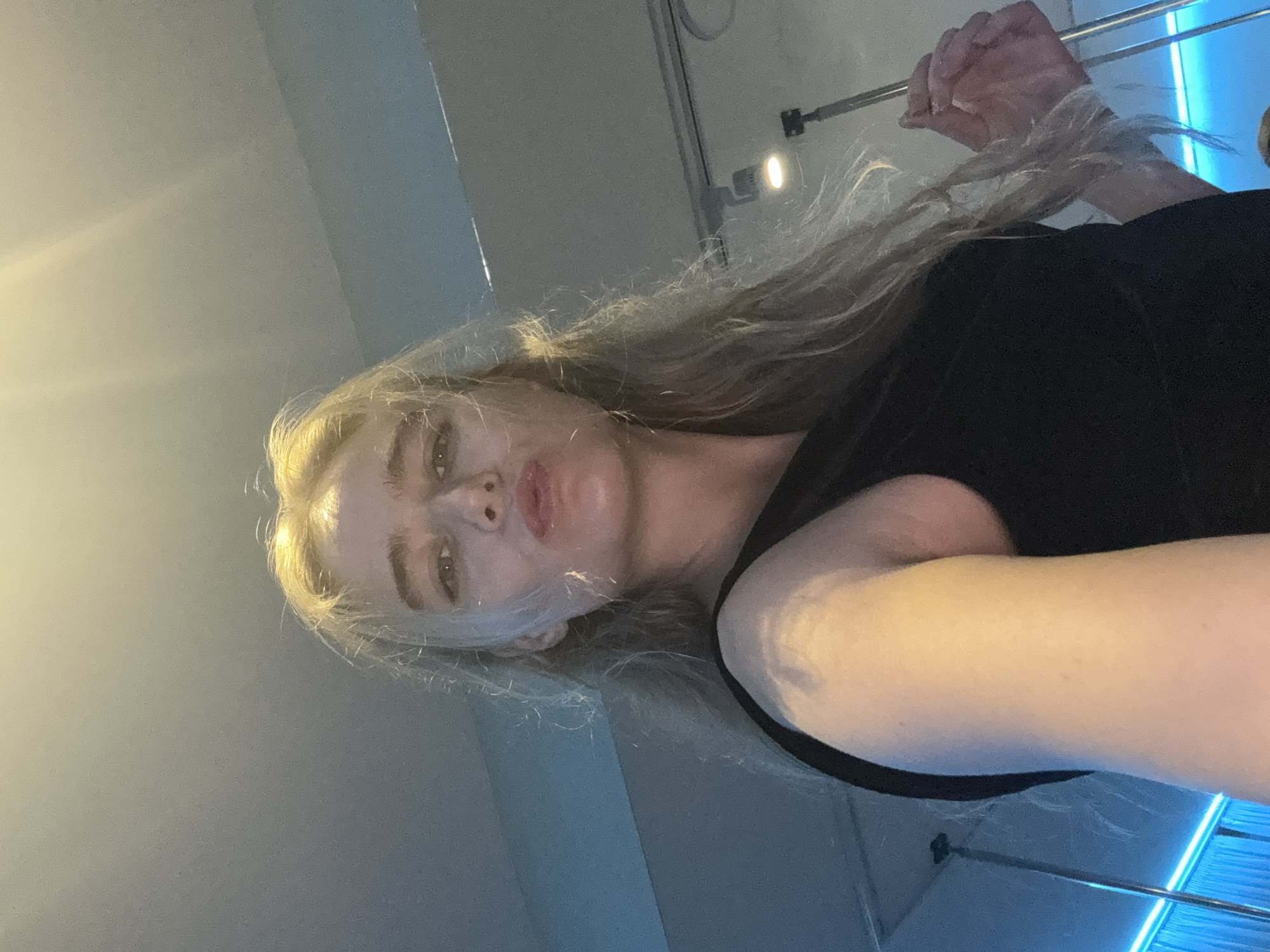 A close-up, tilted selfie of a Caucasian female with blonde hair wearing a black shirt. The location is in a building, city and country unknown. The background includes an off-white ceiling and blue lighting fixtures. The woman is looking at the camera with a neutral expression.