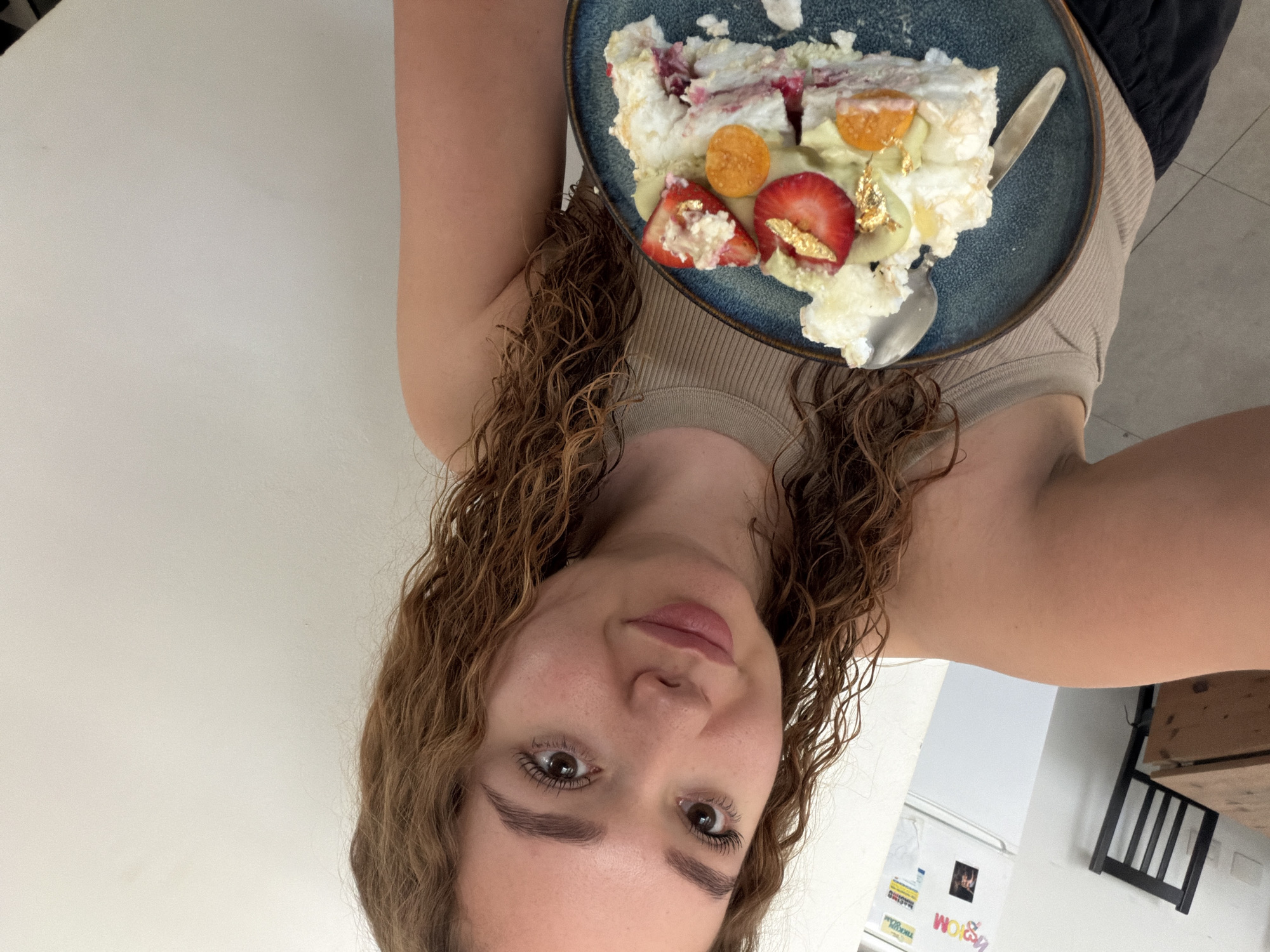 A woman with curly brown hair holds a blue plate with a slice of dessert featuring meringue, fruit (strawberries and other round orange fruits), and what appears to be edible gold leaf. She's wearing a beige tank top. The background shows a white wall, a refrigerator with various magnets and notes, and a black chair. The location is assumed to be an interior space in the city of Ramat Gan, Israel based on the user's request.