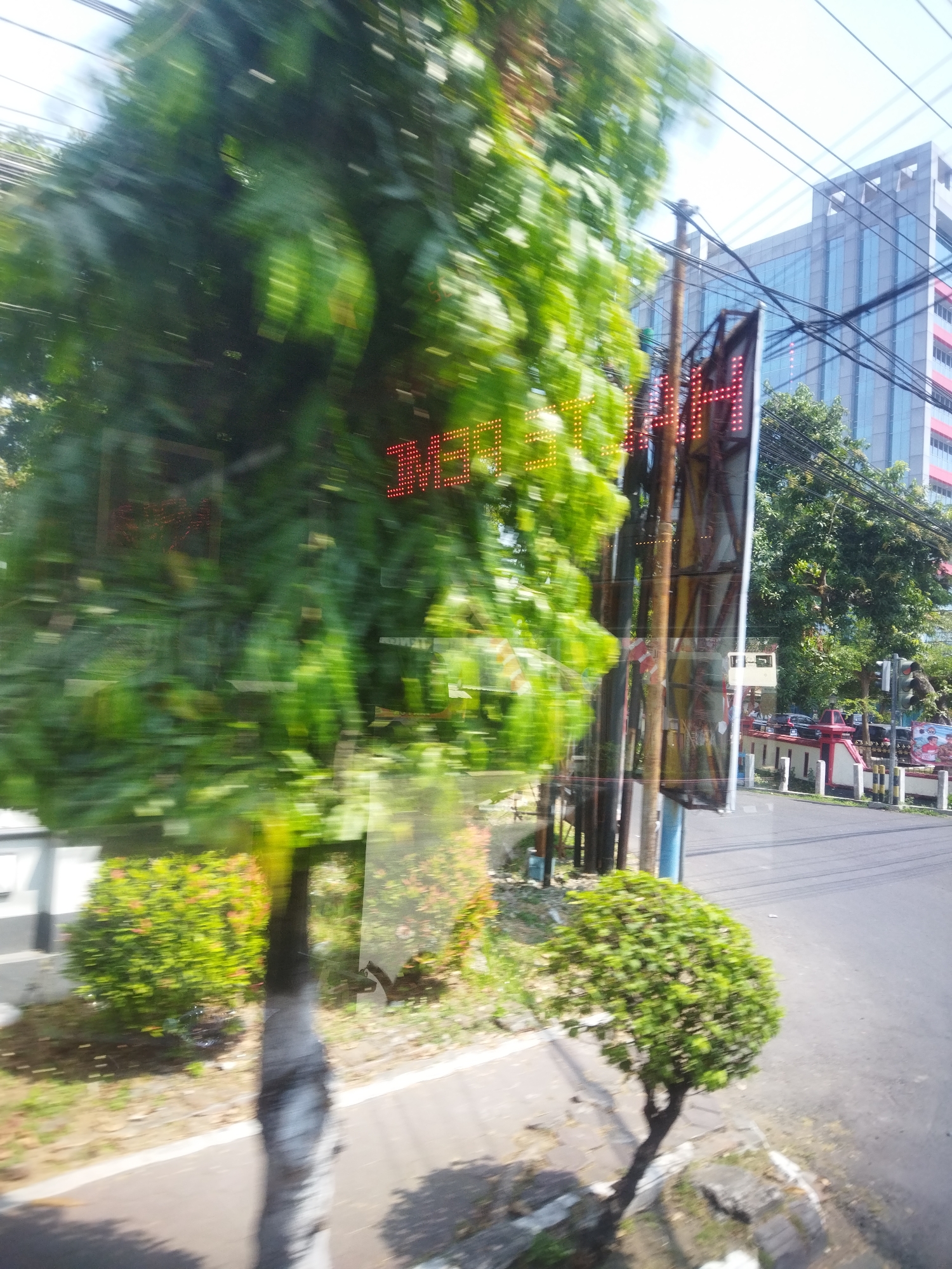 The image depicts a blurry street scene in Jombor, Indonesia, likely captured from a moving vehicle. The foreground is dominated by a large, out-of-focus tree with lush green foliage. To the right, a smaller, more defined tree stands near the edge of an asphalt road. A digital sign with red LED lights is visible, partially obscured by foliage and reflections. In the background, a modern, multi-story building with glass windows stands tall. Electrical wires crisscross the sky above. The street appears to be lined with trees and bushes. A traffic light is visible on the right side of the road. The overall impression is one of motion, with the blur suggesting a fast-moving perspective.