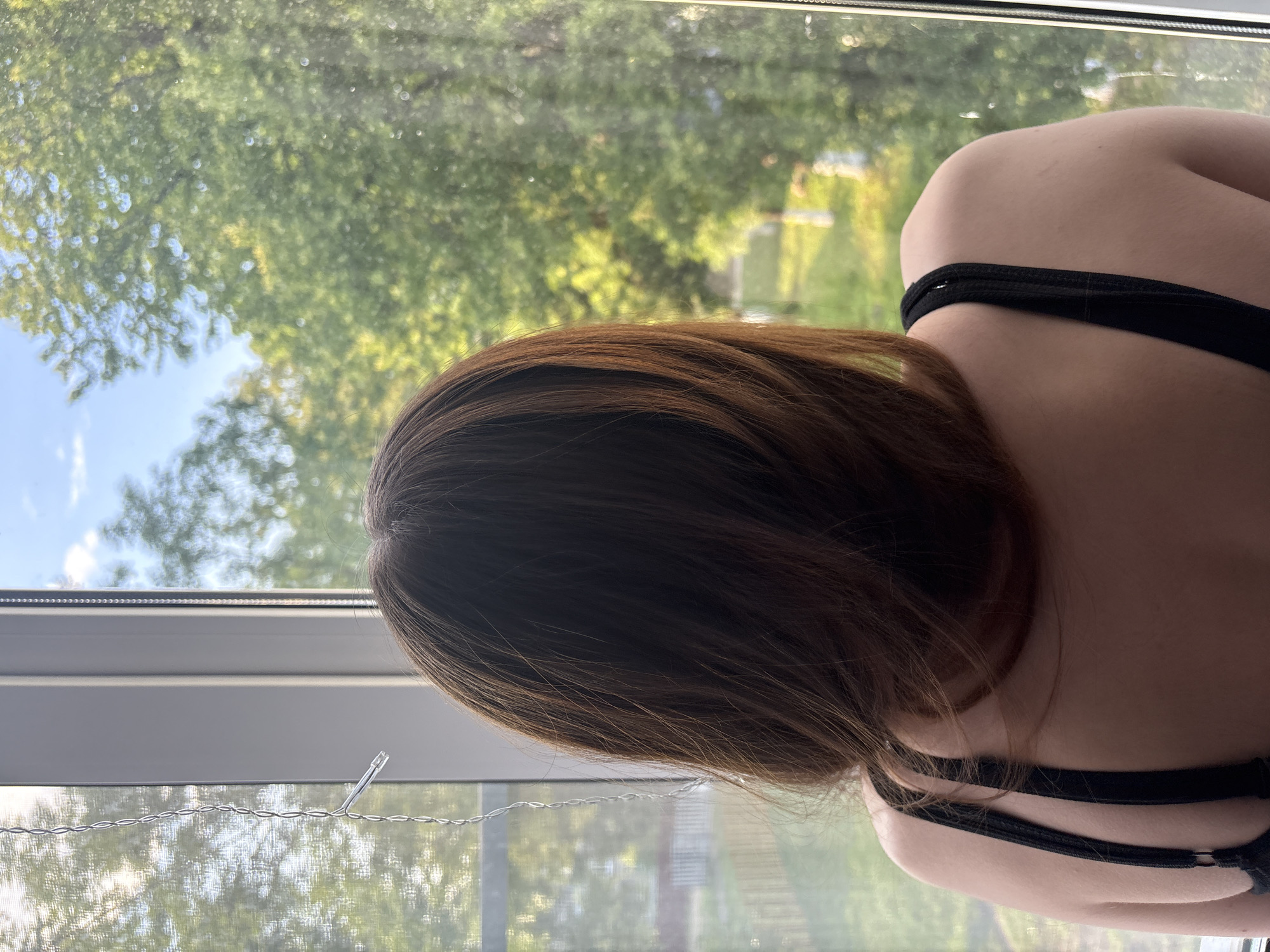 The image shows a close-up view of a person's back and hair, seen from behind. The person is wearing a black bra with thin straps. Their hair is medium-length and appears to be a reddish-brown color. The person is positioned near a window, with their back facing the viewer.
Through the window, a lush green landscape is visible, with trees and foliage bathed in natural light, suggesting it is daytime. The sky appears to be partly cloudy with patches of blue. The window itself has a blind or curtain that is partially visible, and a thin wire is strung across the lower part of the window.
The overall impression is one of a quiet, personal moment, possibly taken indoors looking out at nature. There is no visible text in the image.