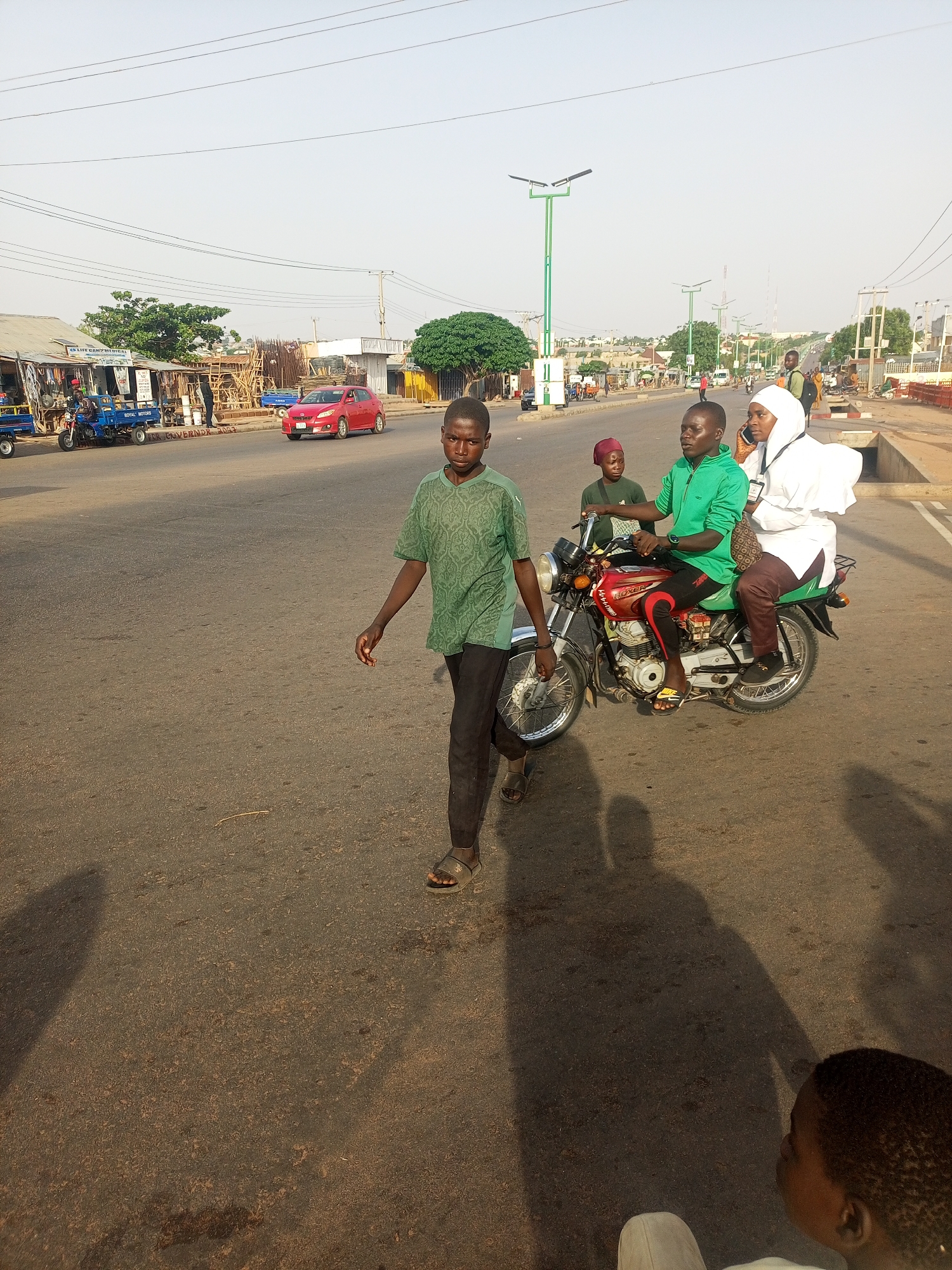 The image captures a street scene in an unspecified location, with a wide asphalt road in the foreground. A young boy in a green patterned shirt and dark pants is walking across the road. To his right, a red motorcycle carries three passengers: a driver in a green jacket, a younger passenger in a maroon head covering behind him, and a woman in a white hijab seated on the back. In the background, buildings and shops line the street, and a red car is visible. Streetlights are spaced along the road. Shadows are cast across the road, suggesting sunlight from the side. A portion of a person's head is visible in the bottom right corner.
