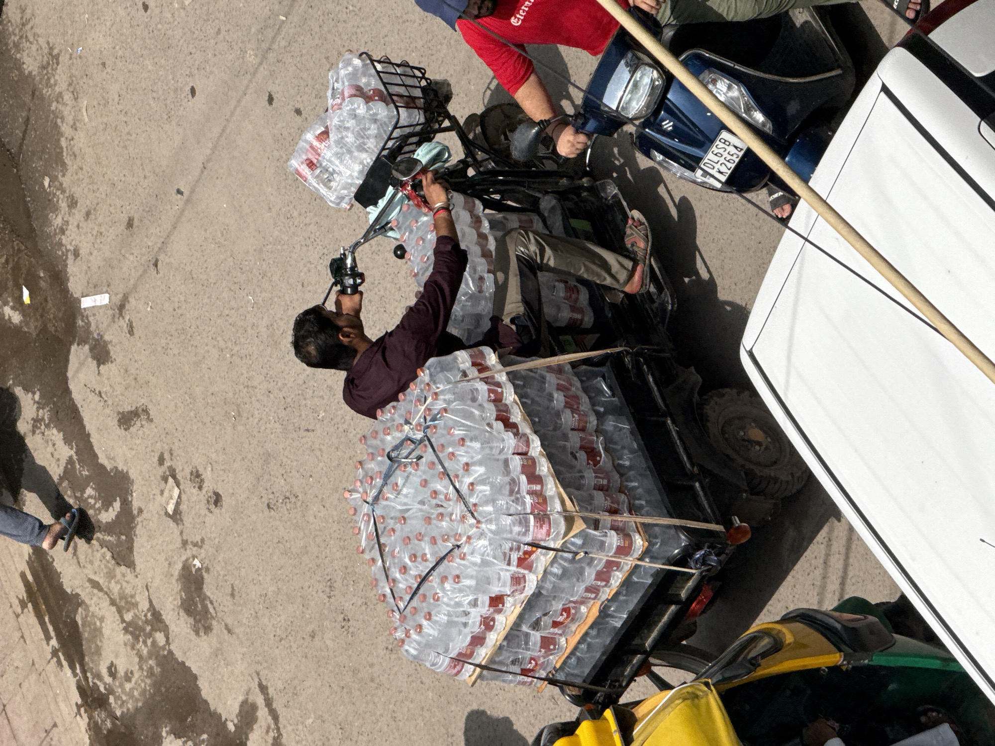 An overhead view shows a street scene in Delhi, India, on a sunny day, with a motor-rickshaw overloaded with bottled water. The rickshaw is a dark color with a flatbed trailer behind the driver. It is stacked high with rows of clear plastic water bottles, each with a red label and a clear or light brown cap. The bottles are secured to the trailer with black straps.
The driver, a man with dark hair, is wearing a maroon shirt and tan pants, and sandals. He is operating the rickshaw, looking ahead and holding onto the handlebars. Next to him, on a dark blue scooter, is another man wearing a red t-shirt and a blue cap. The scooter has a license plate that reads "DL6SB K2654".
In the background, a yellow auto-rickshaw is partially visible on the left. The ground is paved with asphalt and shows some wet patches and debris. The overall scene depicts a typical street view of a commercial area in Delhi, with transportation and daily commerce in motion.