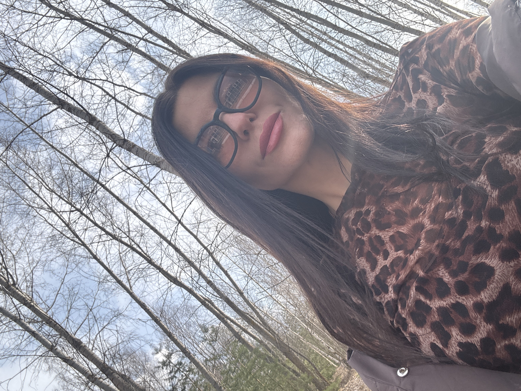 A woman with long, dark hair and glasses is the focal point of a selfie taken outdoors. She is wearing a leopard print shirt, and a grey coat is visible at the edge of the frame. The background reveals a forest of leafless trees against a light blue sky with wispy clouds. Based on the information provided, it is implied the location is in the city of Chaikovsky, Russia.