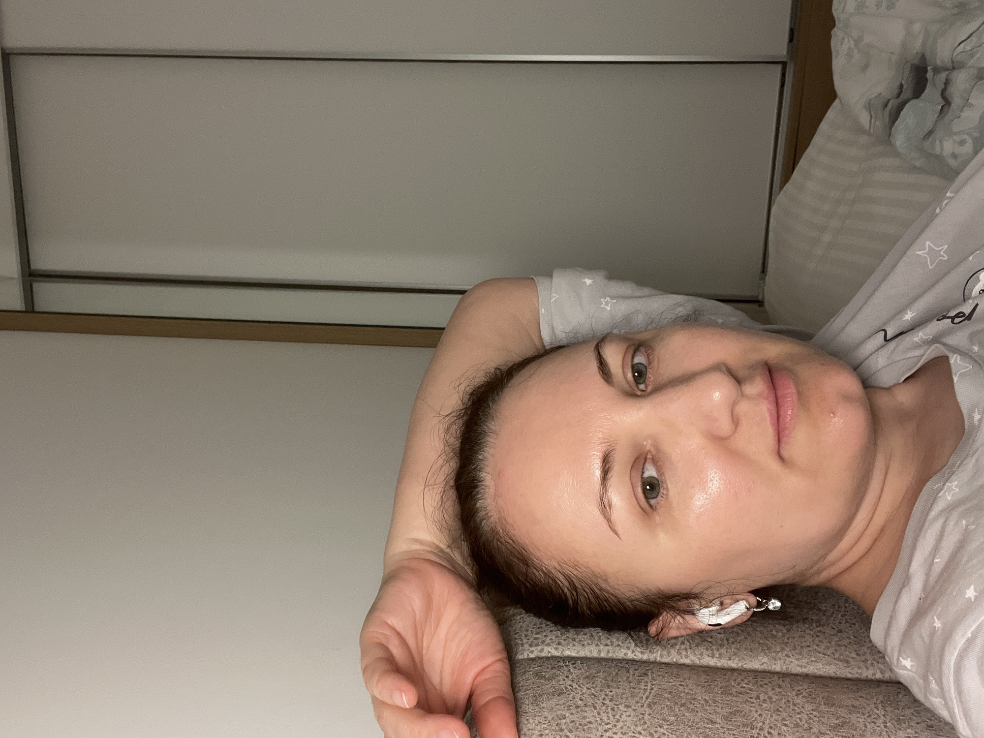 The image captures a woman lying on her left side, looking directly at the camera with a neutral, calm expression. She has dark hair, green/hazel eyes, and her skin appears well-hydrated or dewy. She is wearing a light grey t-shirt patterned with white stars; a faint, partial word, possibly "el," is also visible on the fabric. A small, delicate earring with a clear dangling stone is visible in her right ear. Her left arm is bent, with her hand resting gently near her head. She is propped on a textured, grey upholstered surface, likely the backrest or headboard of a bed or sofa. The background consists of a light grey wall or wardrobe with subtle horizontal and vertical paneling, suggesting sliding doors. Towards the upper right, striped bedding and another grey blanket with white star patterns can be seen. The lighting is soft and ambient, characteristic of an indoor setting, possibly during the evening. This scene is set in Budva, Montenegro.