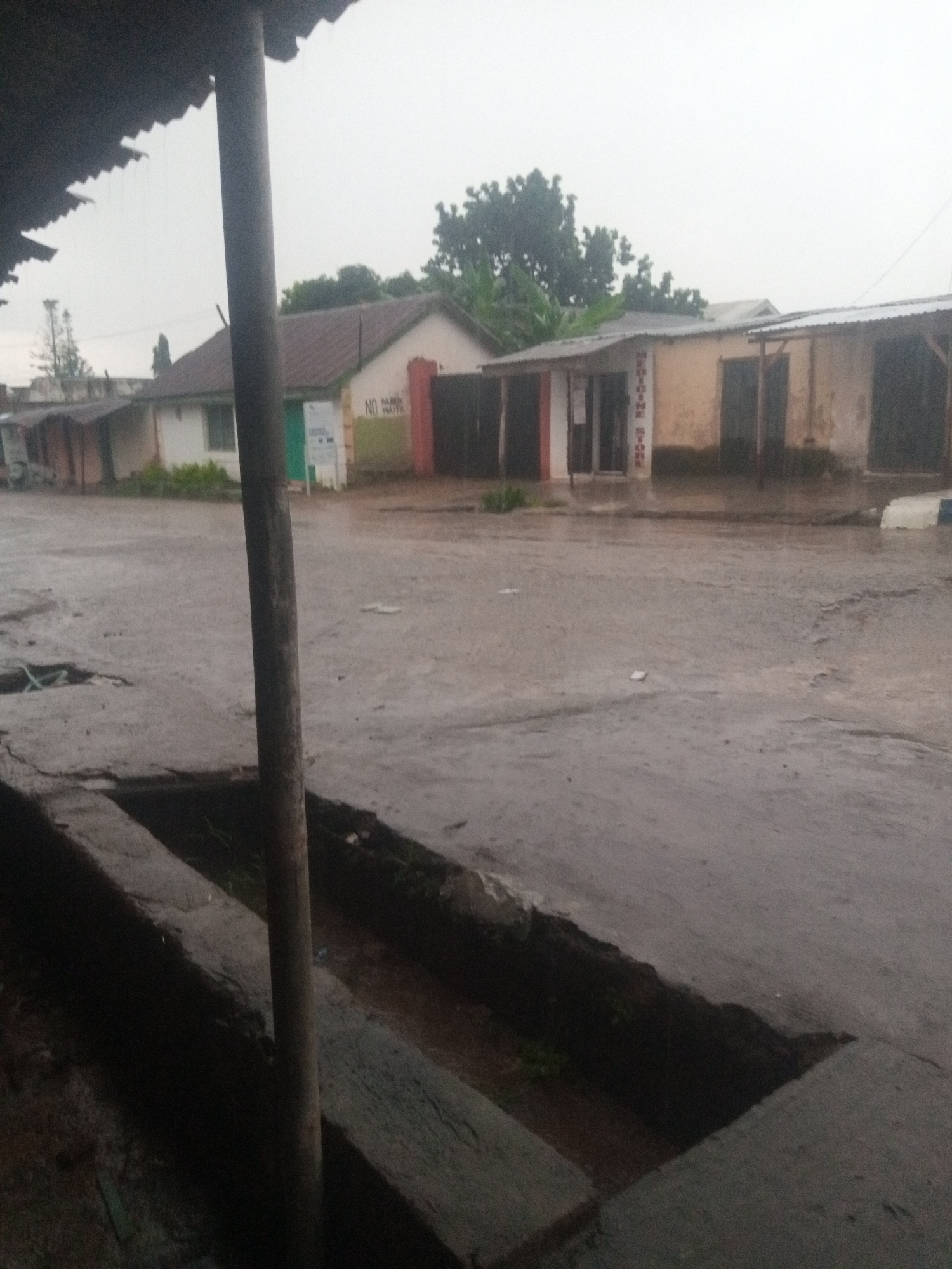 The image depicts a street scene in Jos, Nigeria, likely during a rainy day. The sky is overcast and the ground is wet, indicating recent or ongoing precipitation. In the foreground, a concrete structure, possibly a drain or a raised sidewalk, runs along the edge of the street. A vertical wooden pole stands prominently in the middle ground, casting a shadow on the wet pavement.
Along the street, a row of buildings with corrugated metal roofs are visible. These buildings appear to be small shops or commercial establishments. One building has a sign that reads "MEDICINE STORE" vertically. Another sign on a different building indicates "NO PARKING NO WAITING". The facades of the buildings are weathered, and some have closed doors or shutters. Vegetation, including trees and shrubs, can be seen behind and around the buildings. The overall impression is that of a commercial area in a developing urban environment, experiencing inclement weather. There are no people clearly visible in the image.