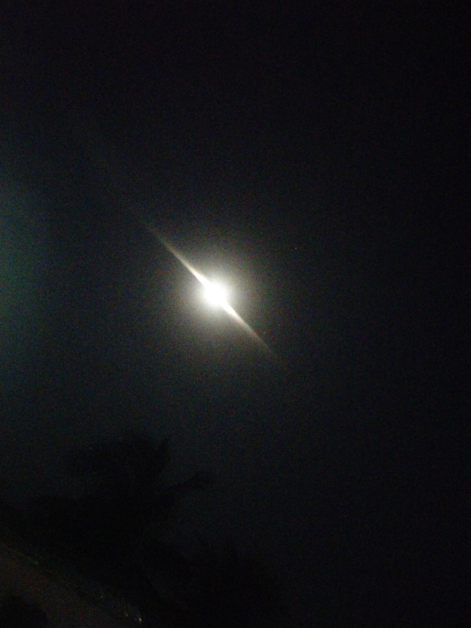 The image depicts a night scene in Unknown, Unknown, dominated by a bright light source, likely the moon or a strong artificial light, with a noticeable glare. The sky appears dark, possibly due to a lack of ambient light or atmospheric conditions. In the lower portion of the image, there's a vague outline of what seems to be a tree or foliage, partially obscuring the view. The scene appears quiet and sparsely populated, emphasizing the light source as the focal point.