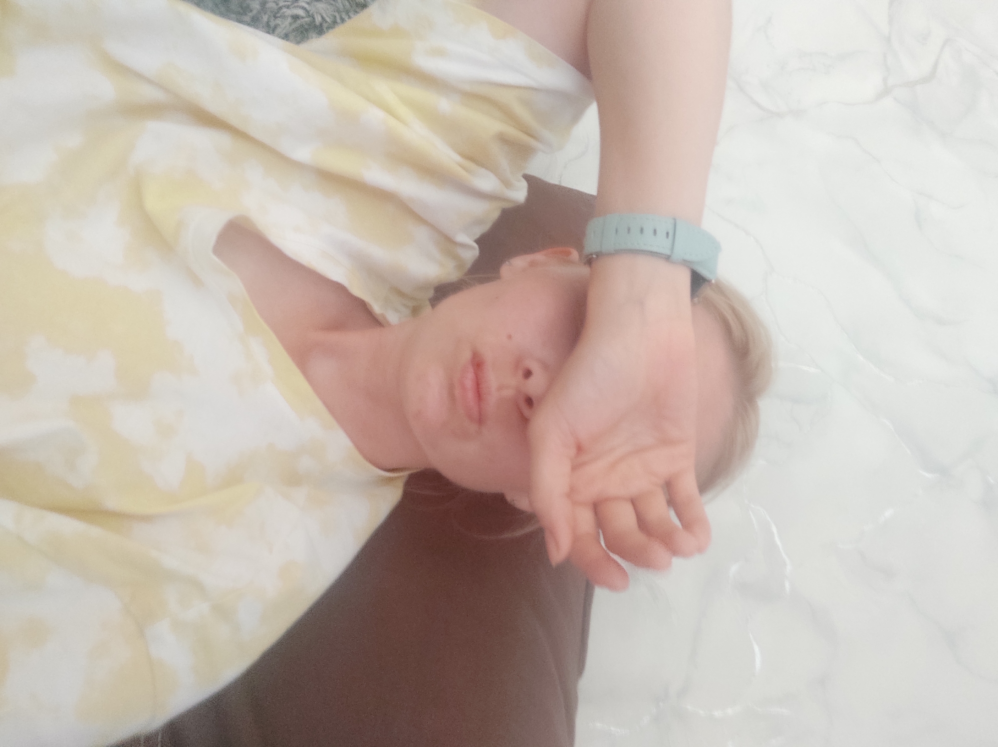A person is lying down, with their face partially obscured by their hand. The person is wearing a yellow and white patterned top. They have a teal-colored wristwatch on their wrist. The person is lying on what appears to be a brown pillow and a white marble-patterned surface. There is no information present in the image to identify the location as Sterlitamak, Russia.