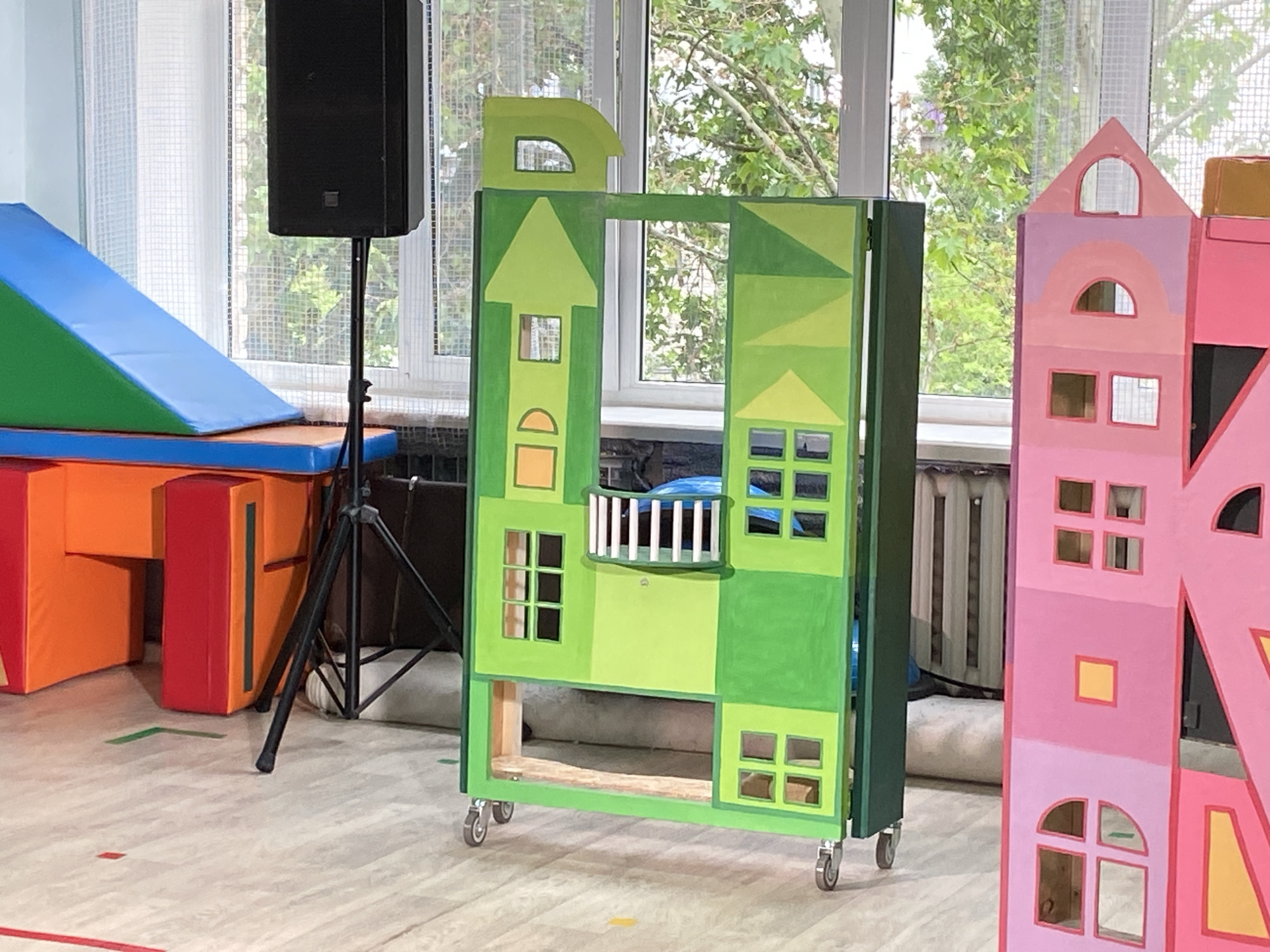 The image captures an indoor scene featuring two colorful, house-shaped structures on wheels, likely props or play equipment.
The prominent structure in the center is green, composed of various shades from light to dark green. It has cutout windows in square and arched shapes, some with white frames. A small white-railed balcony is affixed to its upper section. The top of this structure is shaped somewhat like a stylized letter 'D' or a unique roofline. An opening is present in its center, and it stands on small caster wheels.
To the right, partially visible, is another similar house-shaped structure, painted in shades of pink and purple with yellow accents. Like the green one, it features numerous cutout windows of different shapes.
In the background on the left, there is blue and green soft play equipment, including a ramp and mats, suggesting a play area or gymnasium. A black speaker on a tripod stand is positioned in the mid-ground, near the soft play equipment and behind the green structure. Large windows dominate the background, allowing natural daylight to illuminate the scene and revealing lush green trees outside, indicating a daytime setting. A sheer white curtain hangs on the leftmost window.
The floor is light-colored, possibly wood or linoleum, with colored tape markings including red lines and green, yellow, and red dots. No people are visible, and there is no discernible text in the image. The overall impression is that of a bright, child-friendly environment, possibly a school hall, playroom, or stage set for a performance or educational activity.