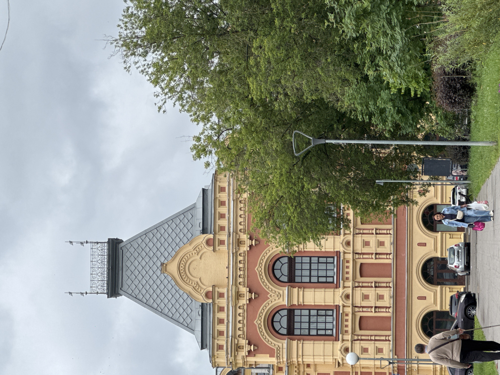 A full shot depicts the exterior of a historical building in Nizhny Novgorod, Russia, on a cloudy day. The building features a prominent mansard roof with grey shingles and ornate yellow and red decorative elements. Two large arched windows with dark frames are visible on the upper floor. In the foreground, a paved walkway runs alongside a grassy area. Two individuals are walking on the path. On the left, a man with grey hair walks away from the camera, using a cane. On the right, a woman walks toward the camera, carrying several shopping bags. Several cars are parked in front of the building's arched entranceways. Lush green trees and bushes surround the building and line the walkway, creating a naturalistic element within the urban landscape.