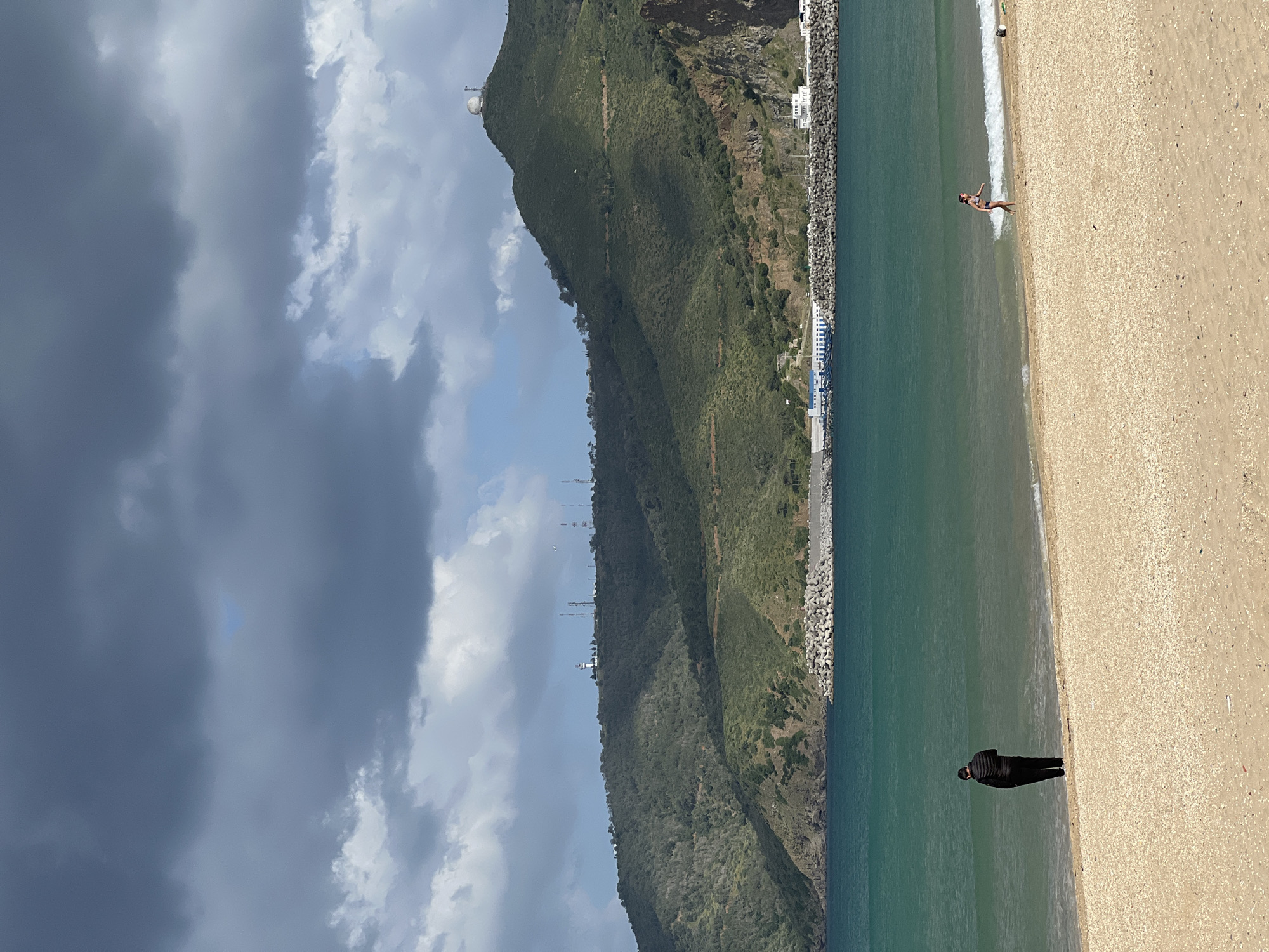 The image captures a beach scene in M'Diq, Morocco. The foreground features a sandy beach, with two figures present. One individual, dressed in dark clothing, stands on the shoreline, facing the water. Further up the beach, another person in swimwear is near the water's edge. The water appears calm and clear. Adjacent to the water is a rocky barrier wall following the shoreline. Behind the wall, a steep, green, mountainous landscape rises. The sky is partly cloudy, with dark, ominous clouds contrasting with patches of blue sky. At the top of the mountain one can see a satellite.