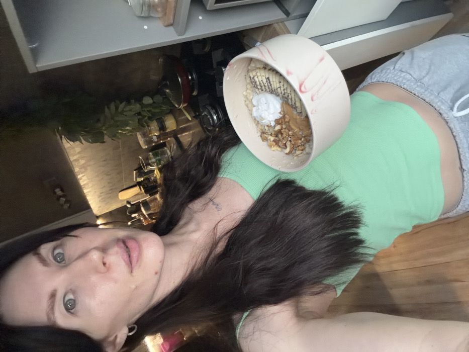 A female individual is captured in a close-up, frontal, waist-up shot within an indoor domestic setting, likely a kitchen. She possesses long, dark brown hair parted on the left, blue-grey eyes, and light skin, with a small silver hoop earring visible in her left ear. A small dark tattoo, appearing to be text, is visible on her upper chest. She wears a light green ribbed tank top that exposes her navel, paired with light grey sweatpants featuring a white drawstring, partially visible at the bottom of the frame. The subject is holding a large, light-colored bowl, possibly ceramic, with reddish-brown swirled patterns, positioned to her left. The bowl contains a prepared meal, identifiable as oatmeal or a similar grain base, topped with sliced bananas, chopped walnuts, a dollop of peanut butter, a dollop of white cream, and sprinkled chia seeds. Her expression is neutral with her mouth slightly open, and she is looking directly into the camera.
The background consists of a kitchen environment featuring a dark-toned backsplash or wall with under-cabinet lighting. On the counter, various items are arranged: multiple glass jars containing spices or ingredients, a red frying pan with a black lid on a black cooktop, a wooden cutting board, and a dark green plant. To the right, a light grey or white cabinet or shelving unit is visible. The lower left portion of the image reveals light brown wooden plank flooring.
The image itself provides no discernible features or contextual clues to confirm the specific location within the city of Perm, Russia, or any city.