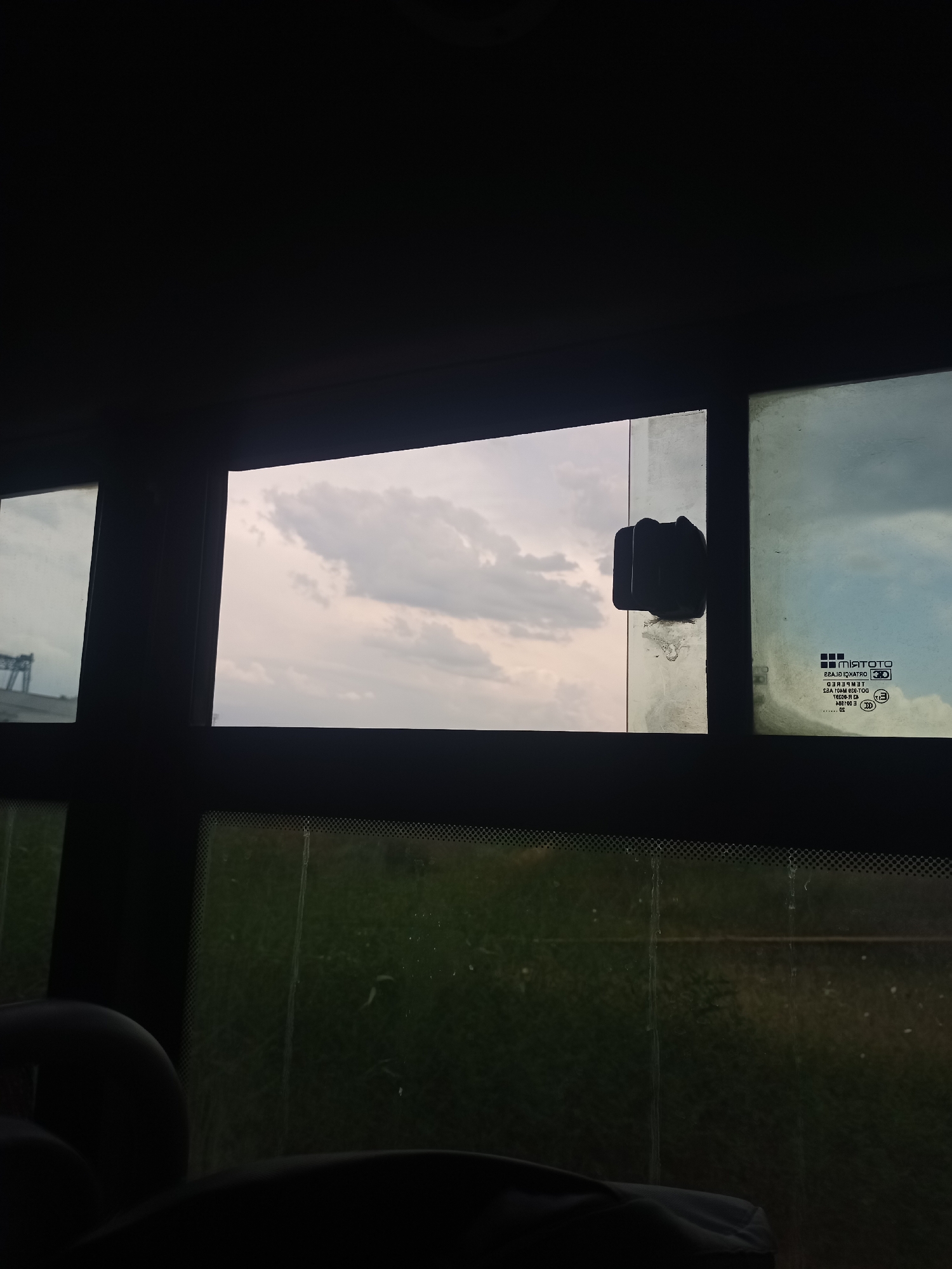 A view from inside a vehicle, looking out through windows at a cloudy sky and green landscape. In the central window, a black suction cup is attached to the glass. The window to the right has a logo and text printed on it, indicating it is tempered glass. In the foreground, dark shapes suggest the interior of the vehicle, possibly seats. The scene appears to be on the outskirts of Boralday, Kazakhstan, given the context provided.
