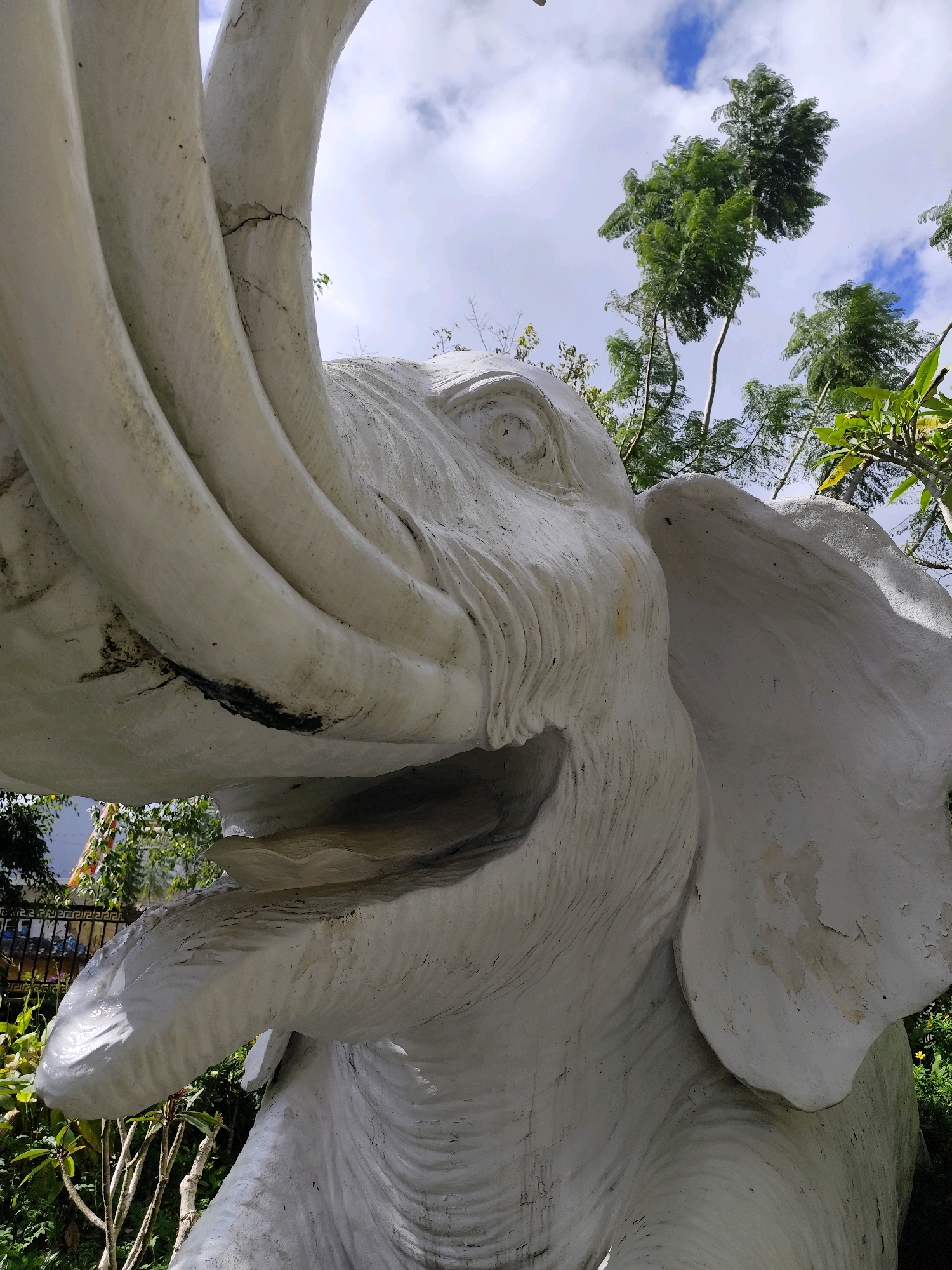 This is a low-angle, close-up, outdoor photograph of a white, weathered elephant statue. The statue is positioned in what appears to be a garden or park setting, with lush green foliage visible in the foreground and background. Above the statue, a partly cloudy sky with patches of blue is visible, along with the tops of green trees. The statue itself is made of concrete or a similar material and shows signs of age, with visible cracks and discoloration on its surface, particularly on the tusks and ears. The detail of the elephant's face, including its eye and wrinkled skin texture, is prominent. The overall impression is that of a large, somewhat neglected, decorative statue set against a natural backdrop. The image is likely taken in Da Lat, Vietnam, a city known for its numerous statues and gardens.