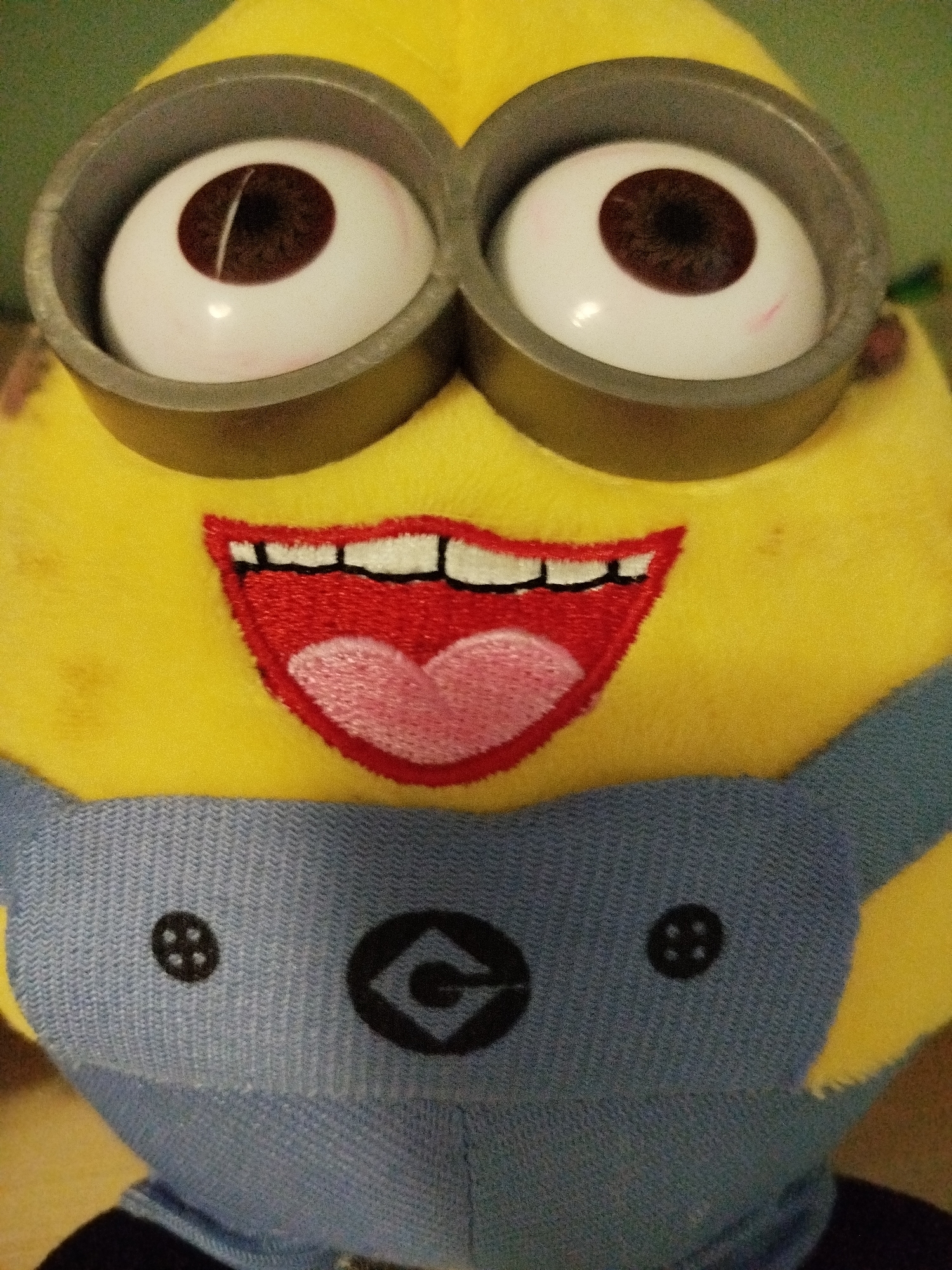 The image is a close-up, eye-level shot of a yellow stuffed Minion toy. The toy has two large, white, bulbous eyes with dark brown irises and black pupils, encased in gray circular goggles. The mouth is embroidered in red with white teeth and a pink tongue. The toy is wearing a light blue, knitted jumpsuit with a black button on each side of the chest, each button featuring a four-dot pattern. A black Minion logo is visible on the chest area. The background is a blurred green surface. The image does not depict a scene in Крывасельскі сельскі Савет, Belarus; it shows a toy in an indoor environment.