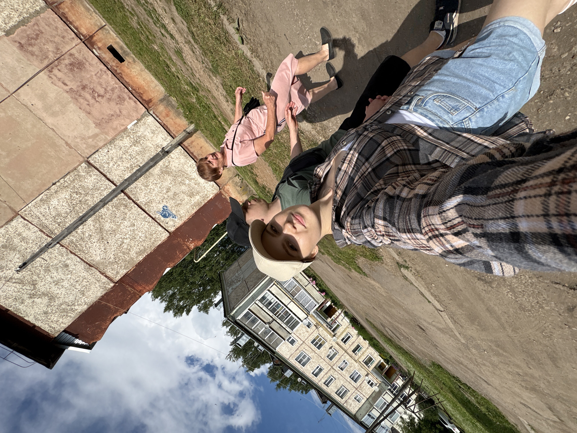 The image shows three people walking outdoors in a residential area. The foremost person, a young woman wearing a plaid shirt and denim shorts, is taking a selfie. Behind her, a young man in a green shirt and a black cap is walking, and further behind, an older woman in a pink dress is walking. They are on a dirt path next to a grassy area and a multi-story apartment building. The apartment building appears to be in the town of Kumyonki, Kirov Oblast, Russia, based on the architecture and style common to post-Soviet era housing. The sky is partly cloudy, and the sun is shining, casting shadows on the ground.