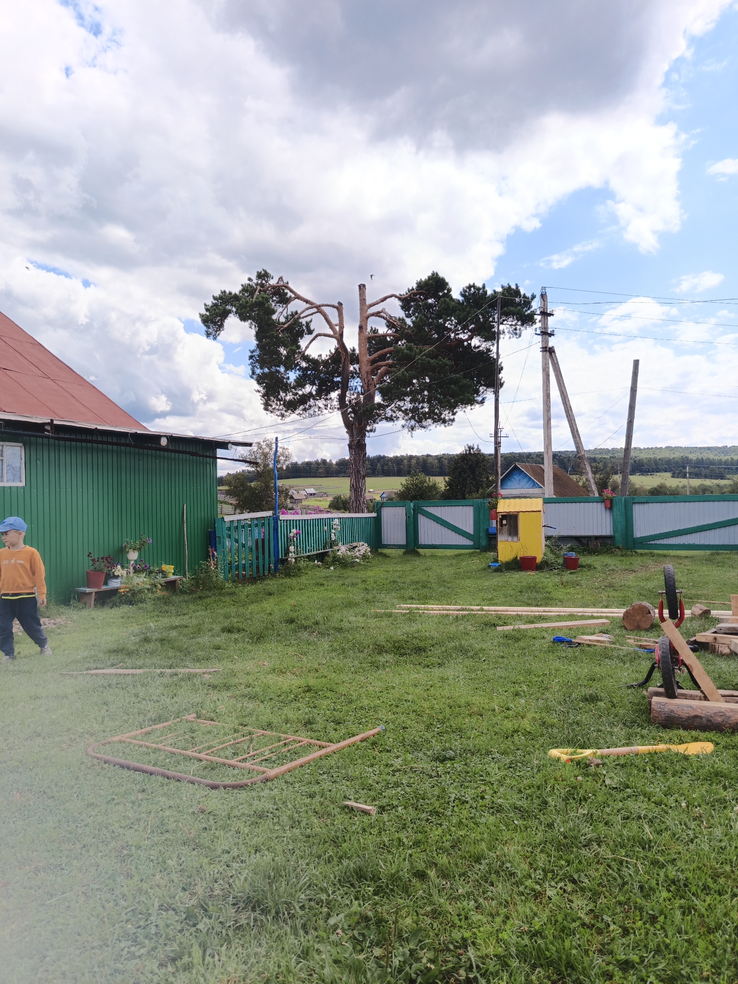 In a rural outdoor setting, likely a village yard in Islambaevo, Russia, a young boy wearing a blue cap and an orange sweatshirt stands on the left, facing the camera. He is walking on a grassy area with scattered building materials. To the left is a green corrugated metal building with a red roof. A turquoise and green fence runs along the middle ground, separating the yard from a further landscape of trees and hills. Behind the fence, a small yellow structure with a blue roof sits in front of a larger building. Various wooden planks, logs, and a wheelbarrow are strewn across the grass in the foreground and mid-ground, suggesting ongoing construction or renovation. A large tree with distinctive branches is prominent in the background, silhouetted against a cloudy sky.