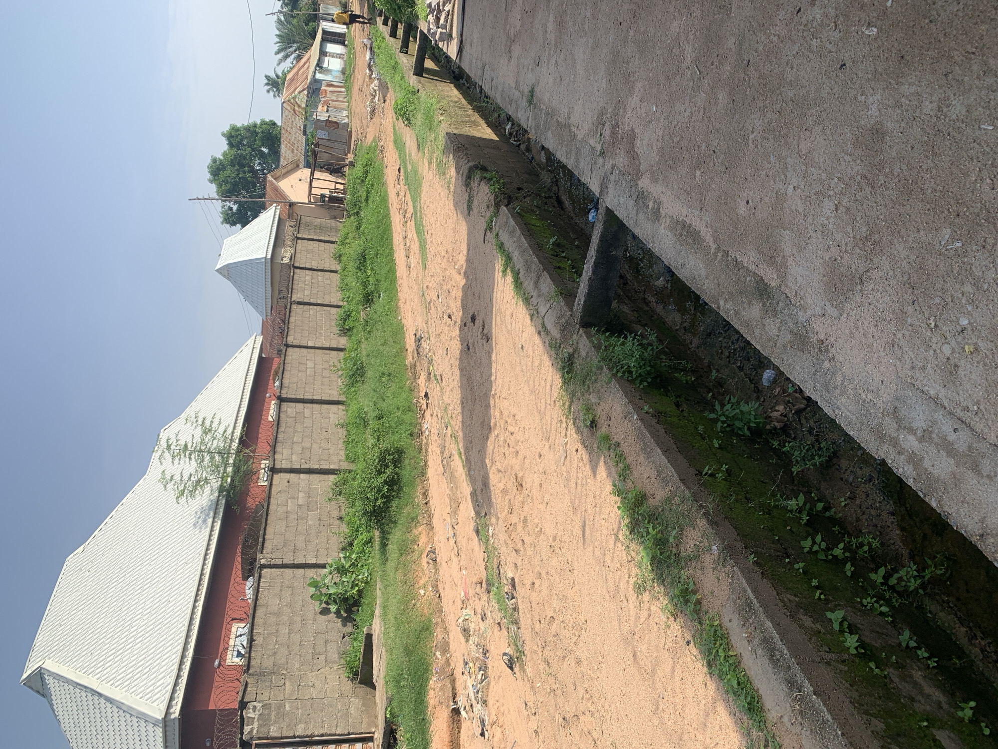 The image depicts an outdoor scene in what appears to be a residential area, possibly in a developing country. In the foreground, a concrete drainage ditch runs along the right side of the frame, filled with murky water and vegetation. The ditch is bordered by a dirt path on the left, which is uneven and scattered with debris. Beyond the path, a low concrete wall topped with barbed wire encloses a property with a red building and a large, white, corrugated metal roof.
Further in the background, a dirt road stretches into the distance, lined with various structures, including small buildings with corrugated metal roofs and some appearing to be shacks. A person in a yellow shirt is walking away from the camera on the right side of the road. The sky is clear and blue, suggesting a sunny day. The overall impression is one of a somewhat impoverished urban or semi-urban environment with basic infrastructure and a degree of disarray.