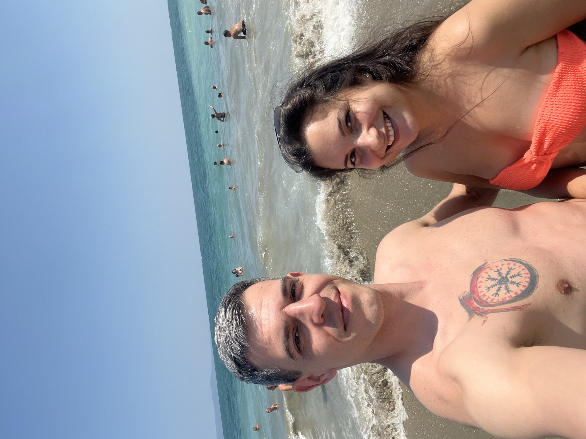 A couple, a man and a woman, are taking a selfie on a sandy beach in Pomorie, Bulgaria. The man, with graying hair and a tattoo on his chest, is on the left side of the frame, smiling. The woman, with dark, long hair and wearing an orange bikini top, is on the right, also smiling. They are both in shallow ocean water with waves breaking behind them. In the background, many other people are swimming and enjoying the sea. The sky is clear and blue.