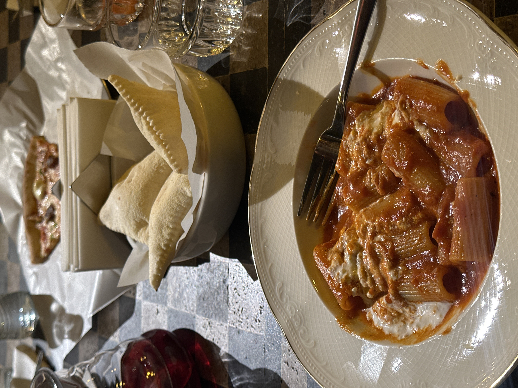 A table setting in what is likely a restaurant features a plate of pasta with red sauce and white cheese, accompanied by a fork. Nearby is a bowl containing flatbread, along with a sandwich or similar item. Glasses of wine and water are also present. The location is likely Yerevan, Armenia, though this is not visually verifiable from the image.