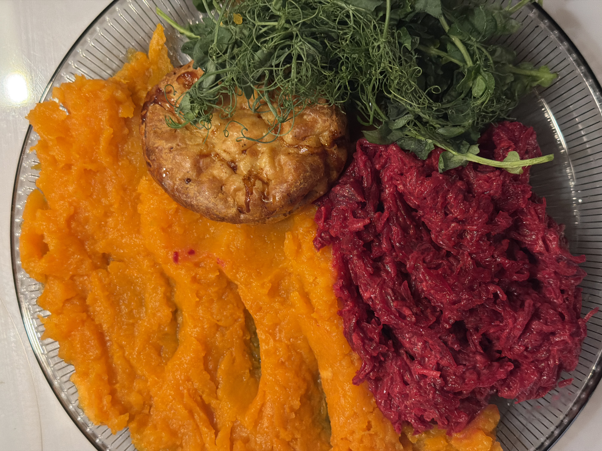 A top-down view of a clear glass plate containing food. A large portion of mashed orange squash or sweet potato occupies the left side of the plate. A circular, golden-brown puff pastry sits atop the squash. To the right, there is a pile of grated red beets. A cluster of green pea shoots is situated at the top, between the pastry and the beets. The plate rests on a white surface. There are no people visible. The exact location in Belgrade, Serbia cannot be determined from this image alone.