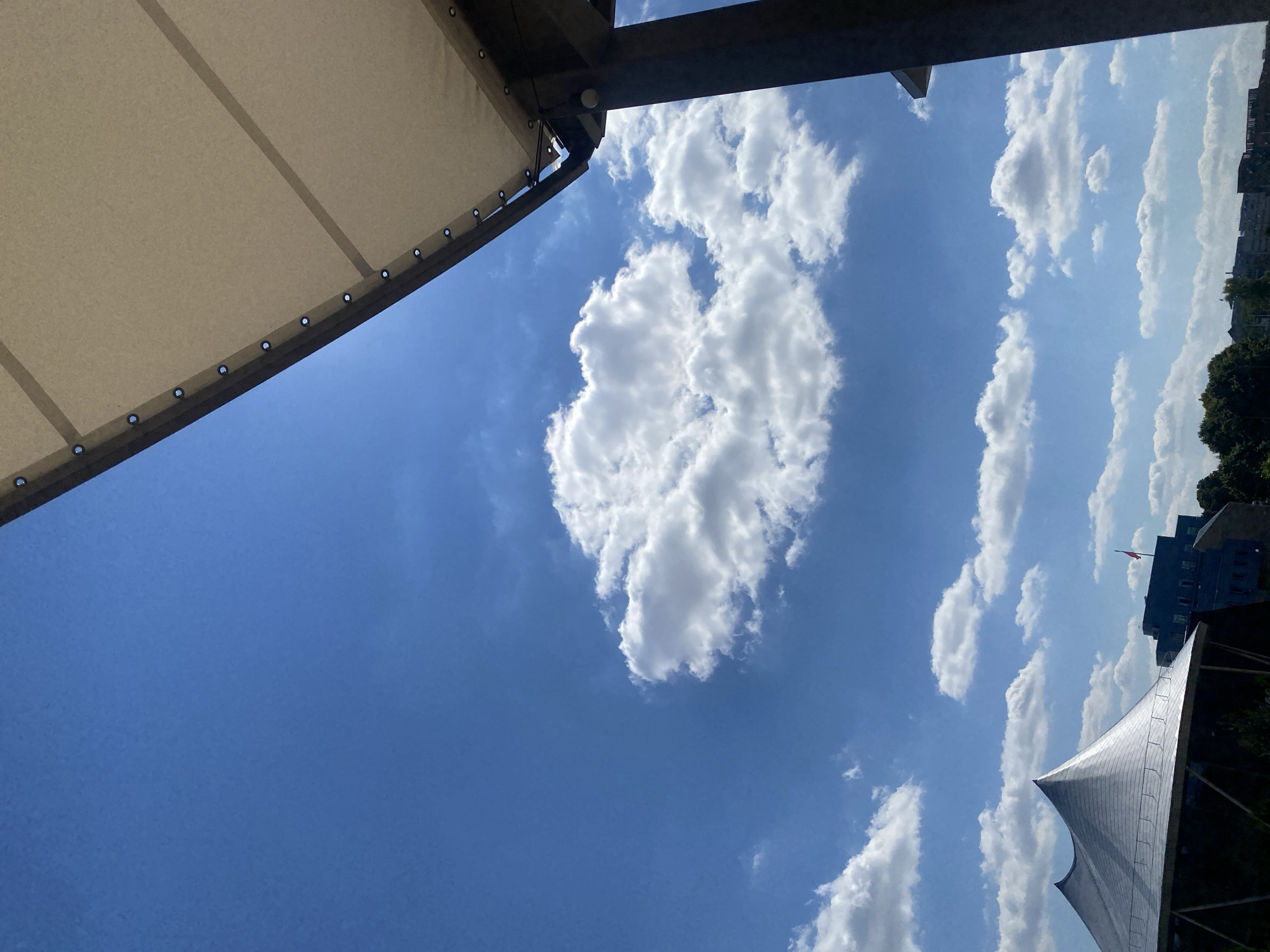 The image depicts a sunny day with a clear blue sky dotted with fluffy white clouds. In the foreground, the edge of a beige awning is visible, supported by dark columns. Below the sky, a large, modern building with a distinctive silver, cone-shaped roof is prominent on the left. To the right of this building, other urban structures and trees can be seen. A red flag flies atop one of the buildings in the distance. The scene suggests an outdoor setting in an urban environment, likely a public space or a commercial area. The composition and elements in the image do not provide specific clues about the location being Minsk, Belarus.