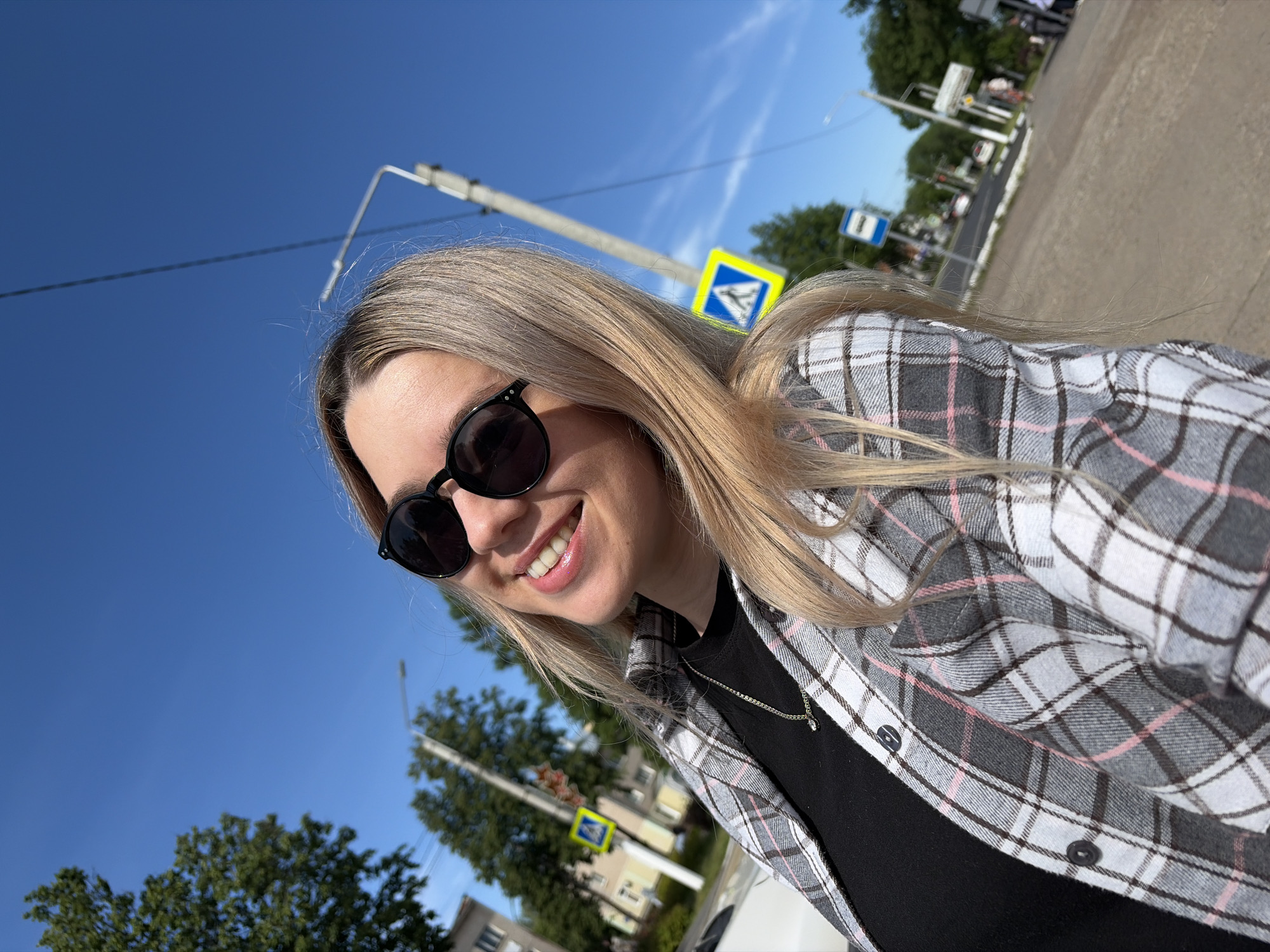 A young woman is captured in a bright, outdoor selfie, likely taken in Chaikovsky, Russia. She is smiling broadly at the camera, wearing dark, round-framed sunglasses, a black t-shirt, and an unbuttoned plaid shirt in shades of grey, white, and pink. Her long, straight blonde hair cascades over her shoulders. A thin, delicate necklace with a small pendant is visible around her neck.
The photo is taken on a clear, sunny day, indicated by the vibrant blue sky and strong lighting. The composition is angled, with the camera positioned low and tilted upwards, showcasing a significant portion of the sky. In the background, typical urban or suburban street elements are visible, including tall utility poles with overhead wires and lush green trees. A yellow diamond-shaped road sign with a pedestrian crossing symbol (an arrow pointing down to a walking figure) is prominently featured on the right side of the image. Further in the blurred distance, what appears to be a paved road or sidewalk, along with other indistinct buildings and vehicles, complete the scene. The woman appears cheerful and relaxed, enjoying the sunny weather.