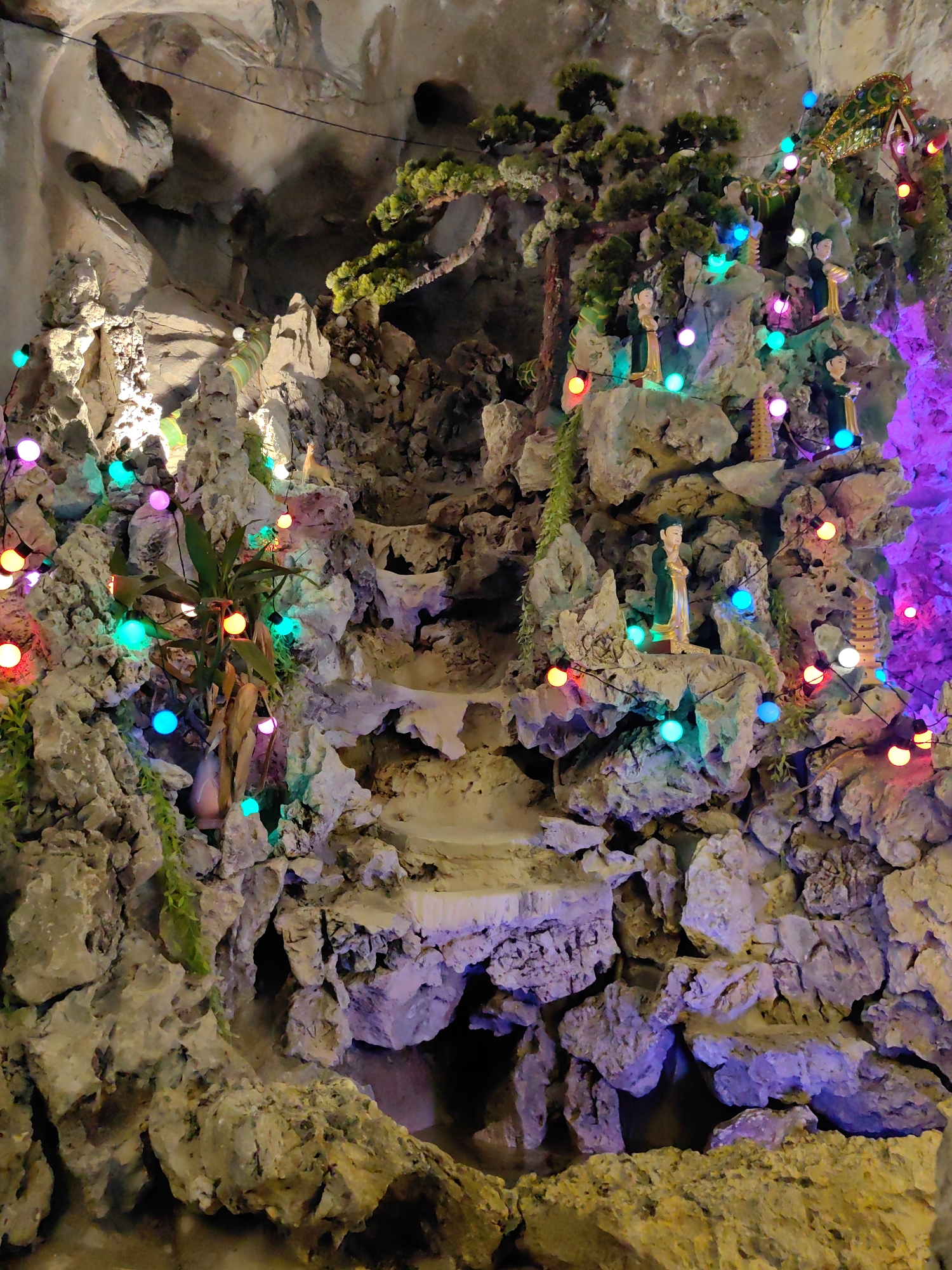 A photograph shows a complex rock formation adorned with figurines, small trees, and multi-colored Christmas lights, located in Lạng Sơn, Vietnam. The rock structure features tiers resembling steps, giving it the appearance of a miniature landscape. Several statues are placed at various points, including figures of people in traditional dress and small pagodas. The overall composition is illuminated with a mix of white spotlighting and the colorful glow of string lights. The background is a continuation of the rock, suggesting the scene is set inside a cave or grotto.