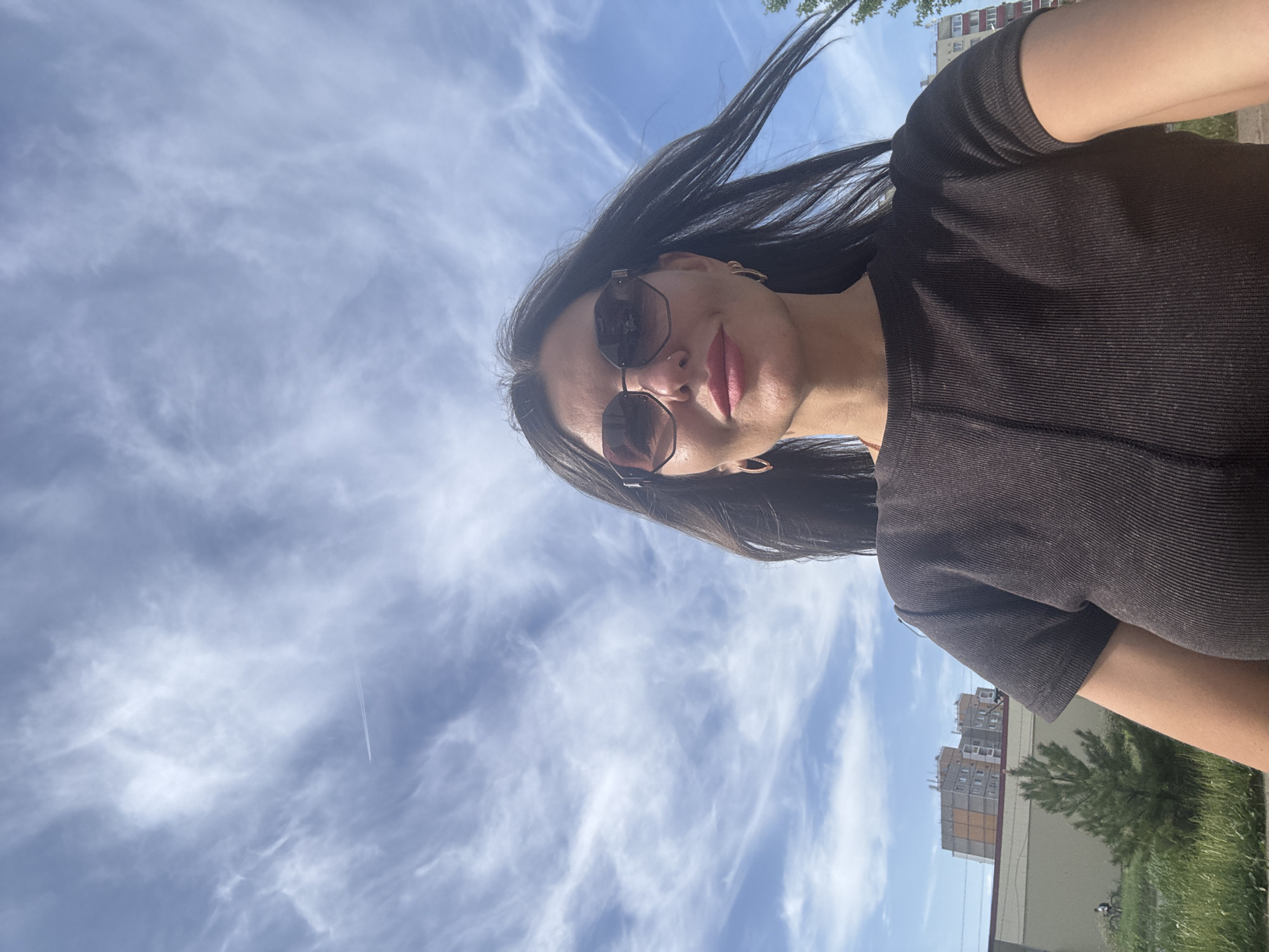 A woman is taking a selfie outdoors with a building and the sky in the background. She is wearing sunglasses and a brown shirt. Her black hair is blowing in the wind. The sky is blue with scattered cirrus clouds. There appears to be a contrail. The buildings are consistent with the architecture of Chaikovsky, Russia. It is daytime.