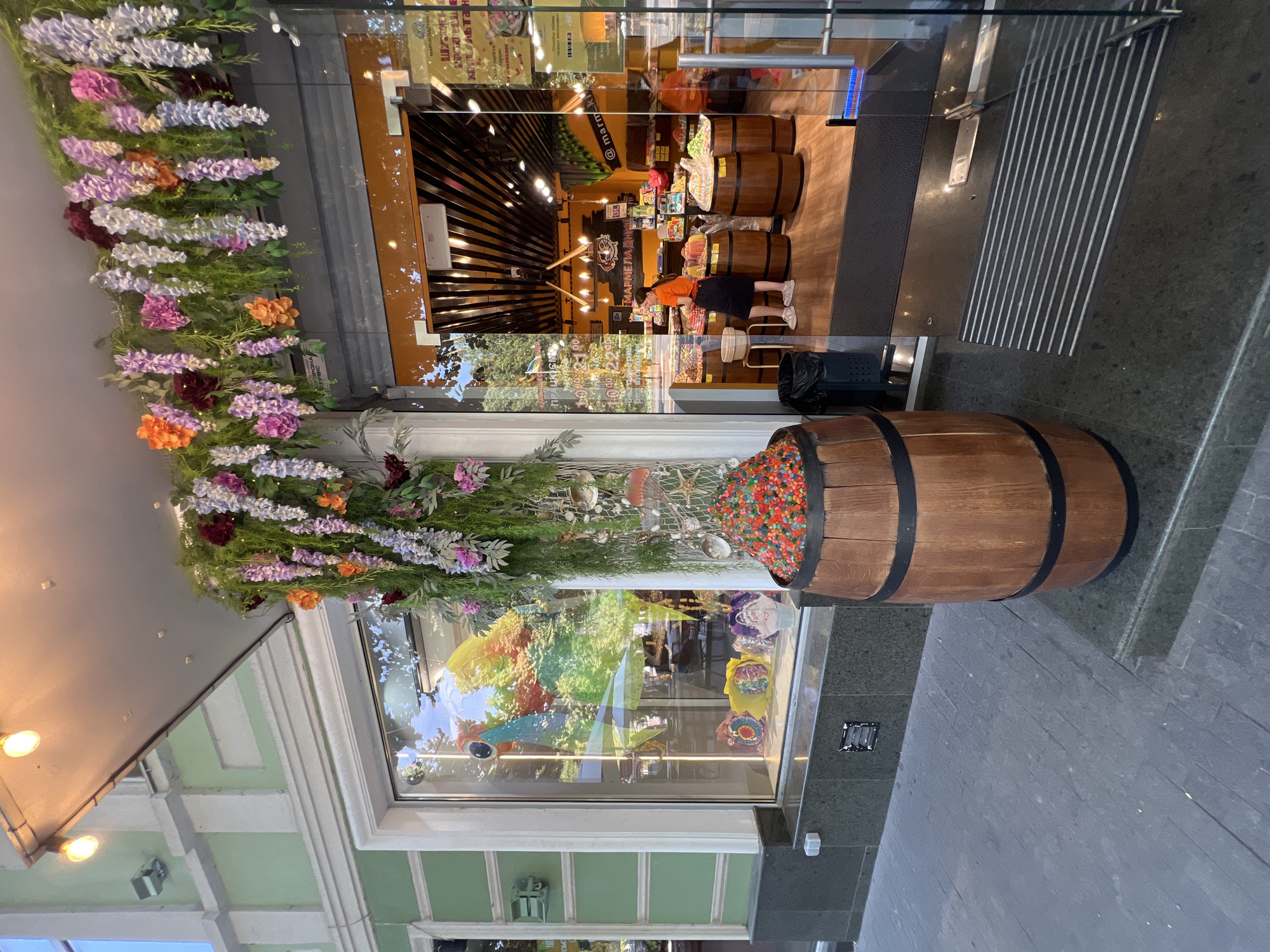 The image depicts the exterior of a confectionery store located on a street in Odesa, Ukraine. The store's entrance features a glass door and a large display window. Hanging above the entrance is an elaborate floral arrangement of artificial wisteria and other flowers in shades of purple, pink, and orange, intertwined with greenery.
In front of the store, to the left of the entrance, is a large wooden barrel overflowing with colorful jelly candies. The display window on the left showcases a vibrant, rainbow-colored parrot decoration, with several gift bags visible inside.
The store interior, visible through the glass entrance, is brightly lit and decorated with orange and black accents. Wooden barrels, similar to the one outside, are used as displays for more candies. A young woman wearing an orange shirt and a dark skirt is standing inside, interacting with the merchandise. Various candy products are arranged on shelves and in the barrels.
The street in front of the store is paved with dark stone tiles. The building facade is painted a light green with white architectural details. The scene suggests a warm, possibly sunny day, given the bright lighting and reflections in the glass.