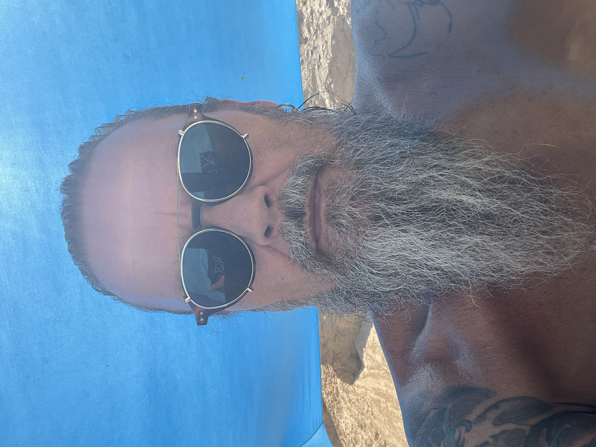 A man with a long grey beard and tattoos on his shoulder is looking at the camera while wearing sunglasses. He appears to be outdoors in a sandy area with a blue tarp above him, suggesting a beach or picnic setting. The image was taken in Ga'ash, Israel.